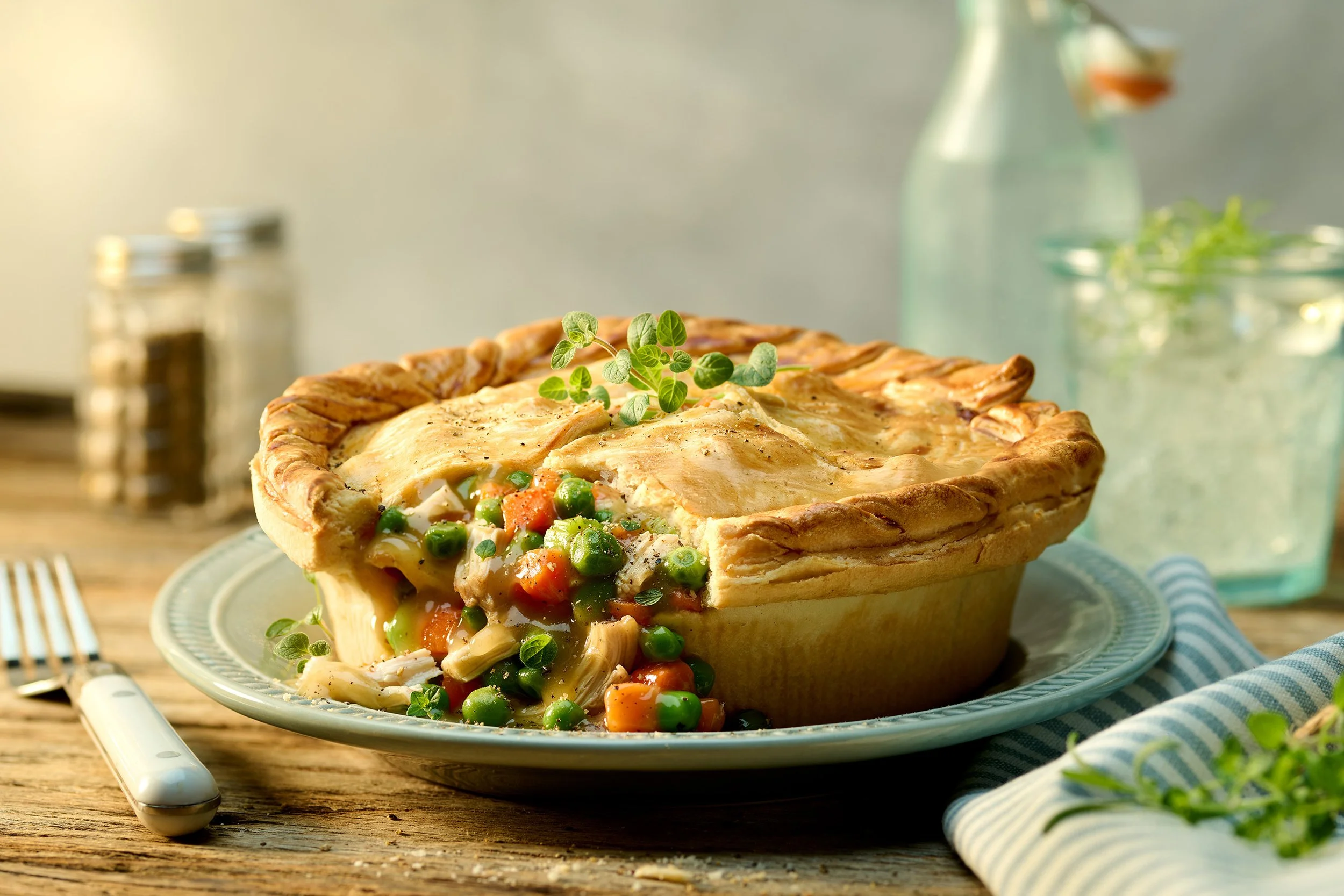 A chicken pot pie with a golden crust, garnished with herbs, with a side of mixed vegetables spilling out from a slice, on a decorative plate; background includes jars and a glass bottle.