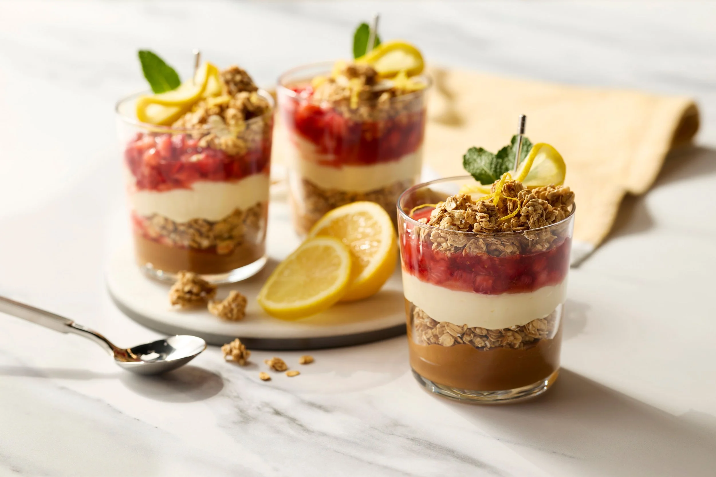 Three layered parfait desserts in glasses with granola, cream, fruit compote, and lemon slices on a white surface.