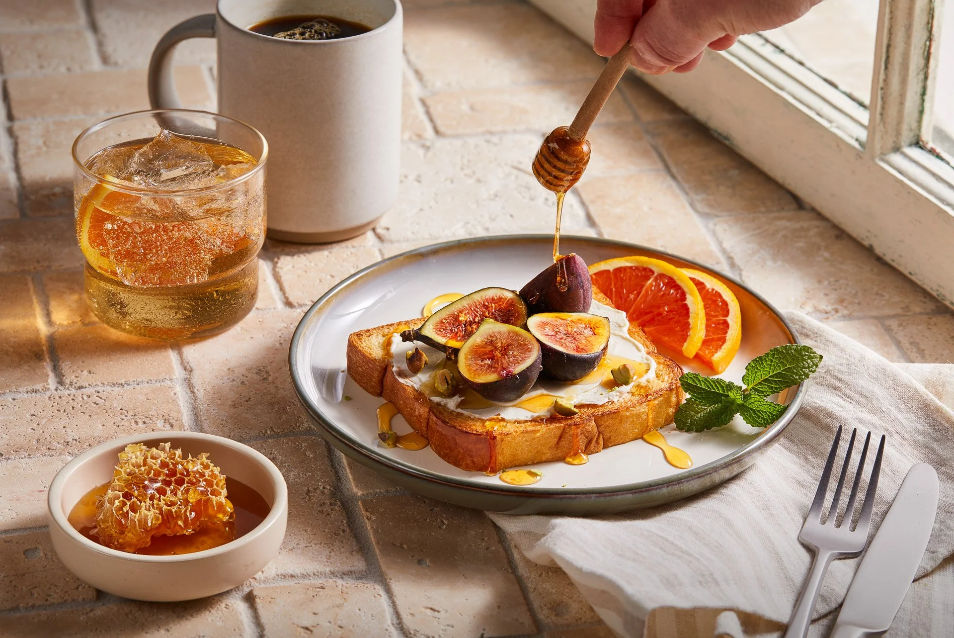 A hand drizzling honey over toast with figs, cream cheese, and pistachios on a plate with orange slices and mint. Nearby are a glass of iced tea, a mug of coffee, and a dish of honeycomb.