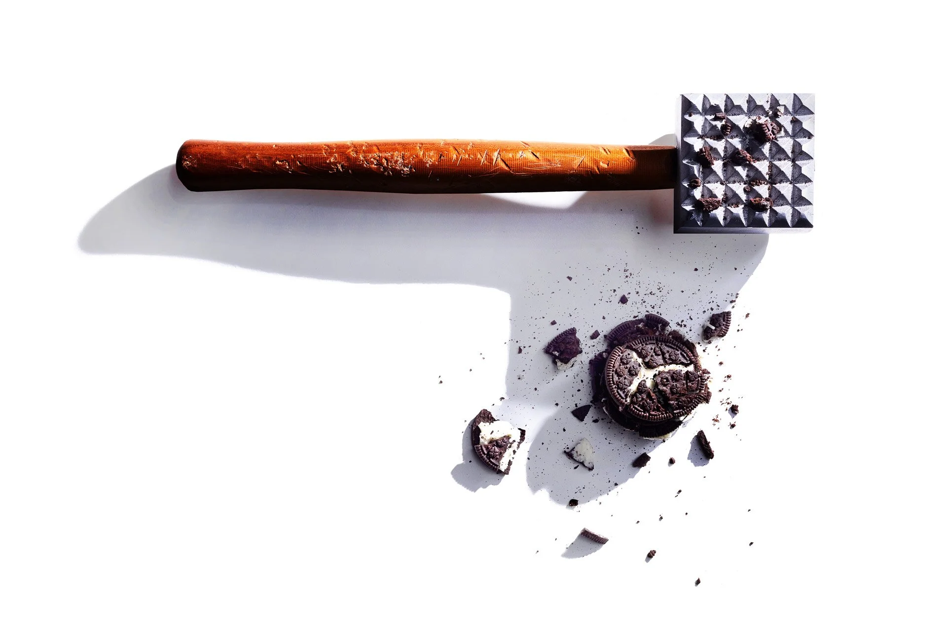 Meat tenderizer crushing a chocolate sandwich cookie on a white background, with cookie crumbs scattered.
