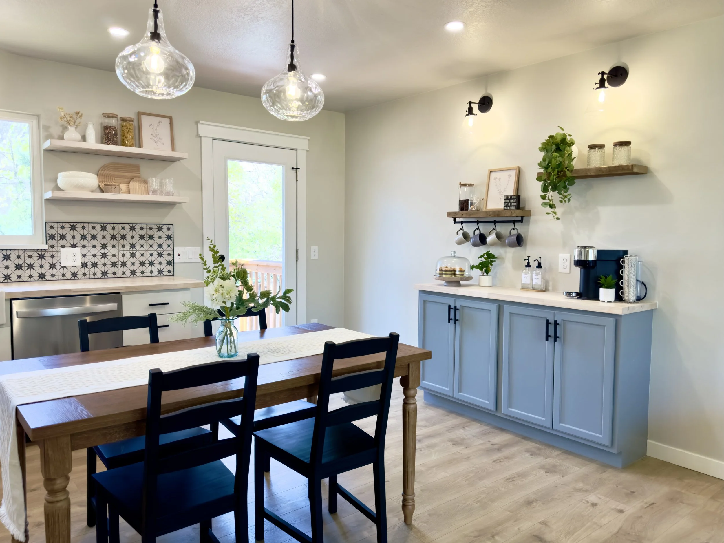 Open kitchen and dining area with a wooden table, black chairs, a blue cabinet, floating shelves with decor, pendant and wall lights, and a door leading outside.
