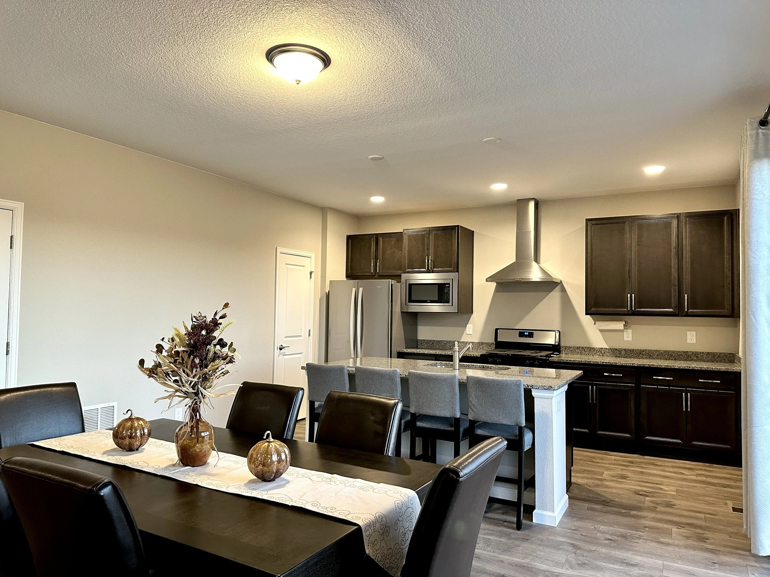 Modern kitchen and dining area with dark wood cabinets, gray bar stools, granite countertops, a stainless steel fridge, microwave, oven, and a dining table with black chairs and a centerpiece of fall-themed decor.