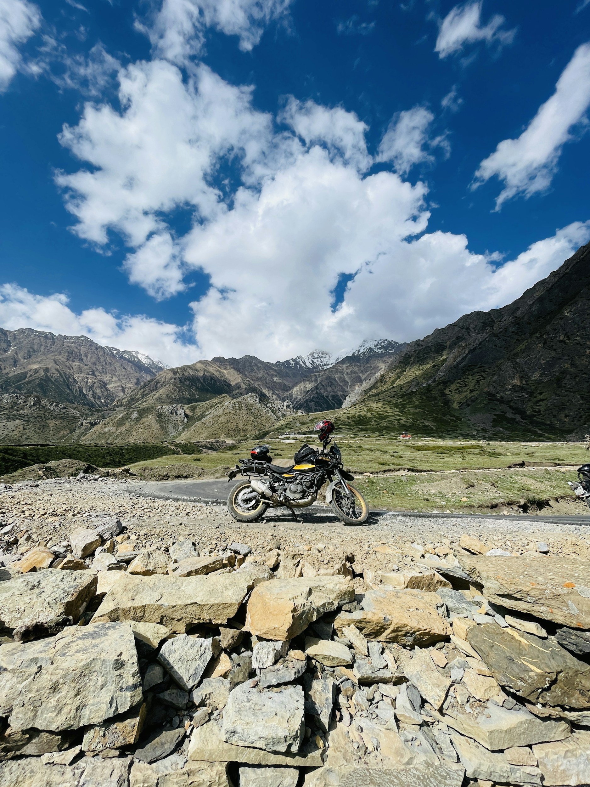 A motorcycle parked on a winding mountain road with rocky terrain, green hills, and snow-capped mountains in the background under a partly cloudy blue sky.