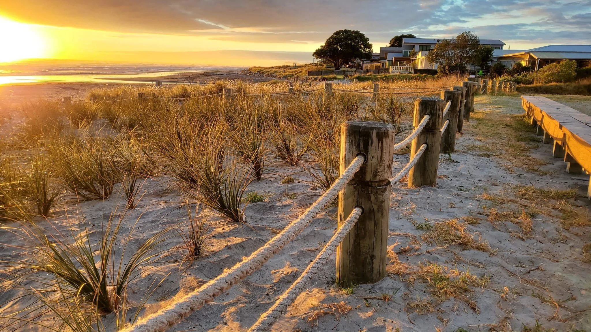 A sandy beach pathway lined with wooden posts and rope railing, leading toward houses and trees in the distance, with the sun setting over the ocean horizon.
