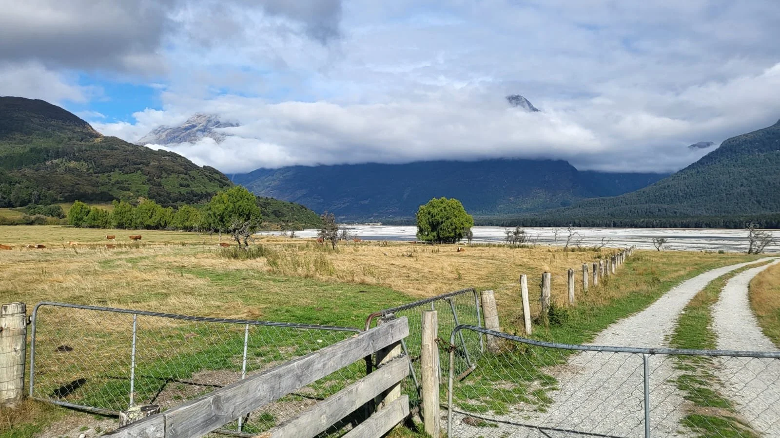A scenic rural landscape with a dirt road, green fields, trees, and mountains in the background, partially covered with clouds.