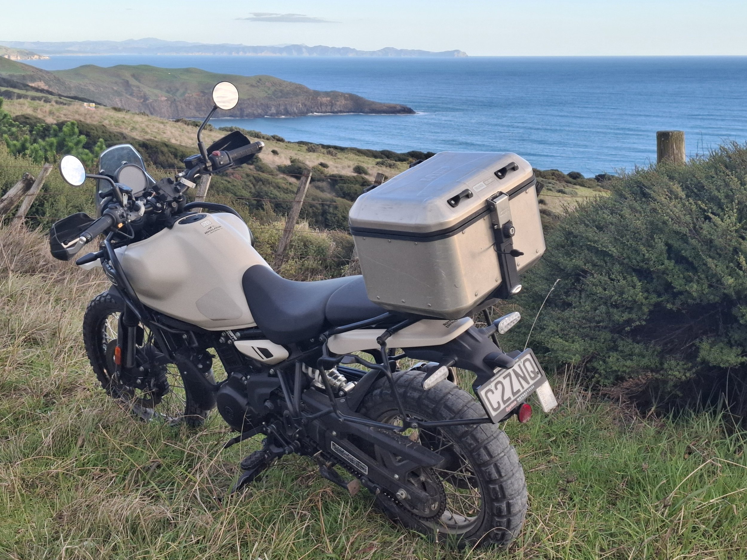 A white motorcycle parked on grassy terrain overlooking the ocean and rolling hills in the distance.