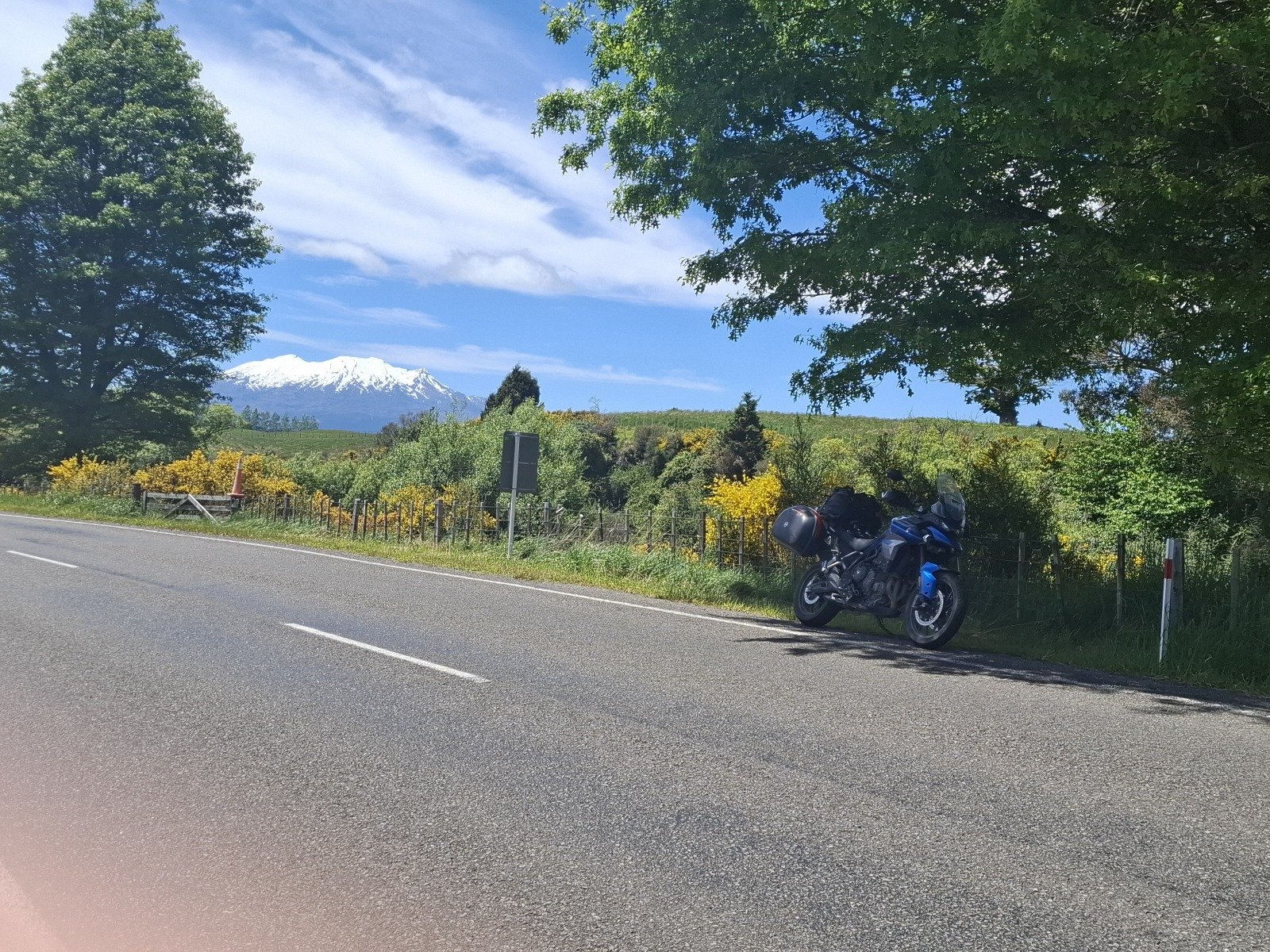 A blue motorcycle parked on the side of a two-lane road surrounded by green trees, with a snow-capped mountain in the distance under a partly cloudy sky.