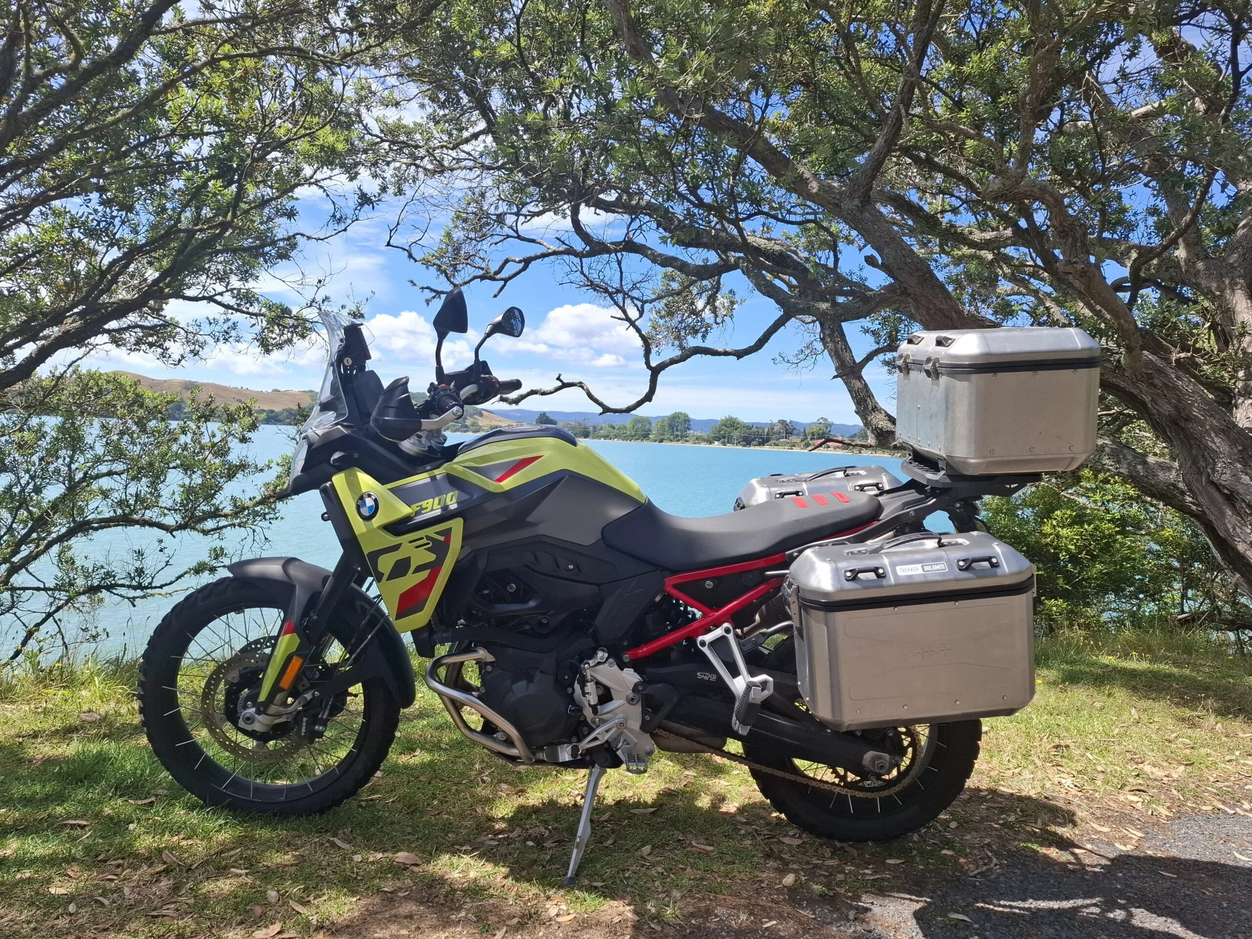 A BMW F900 motorcycle parked on grass beside a lake, with trees and sky in the background.