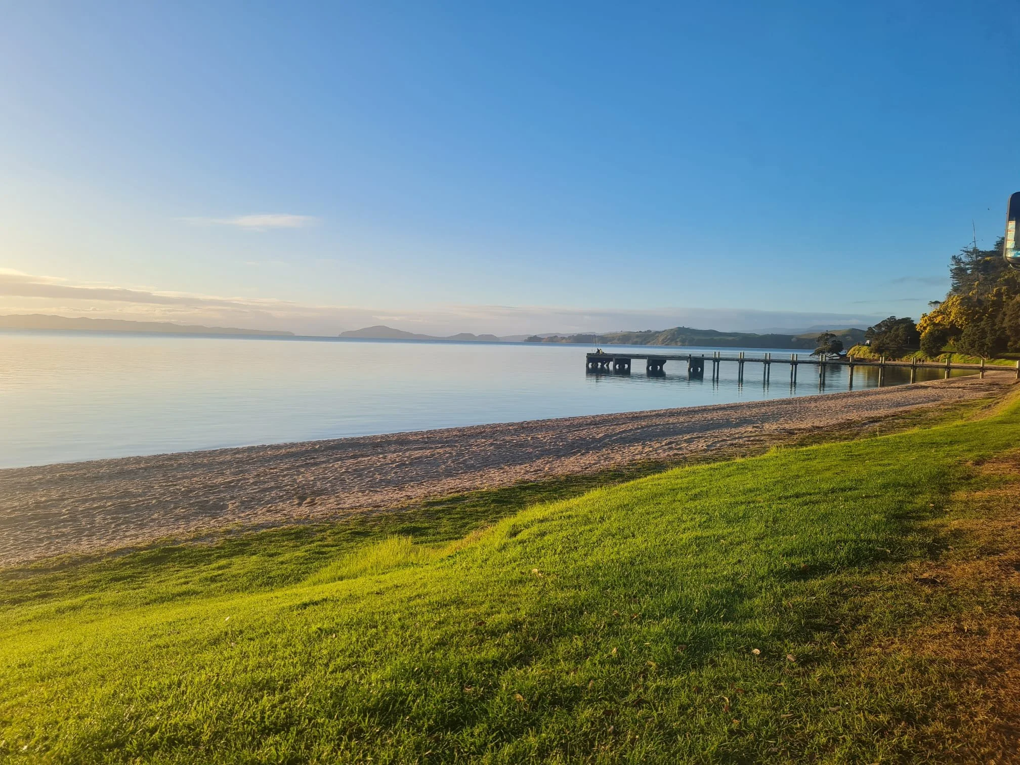 Sunset over calm water with a pier extending into the harbour, sandy beach, and grassy area with trees on the right.