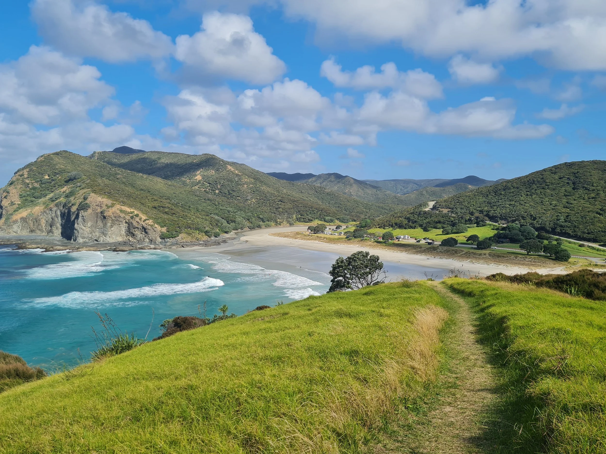 A scenic coastal landscape with a grassy hillside path leading toward a beach, with ocean waves, green hills, and a partly cloudy sky in the background.