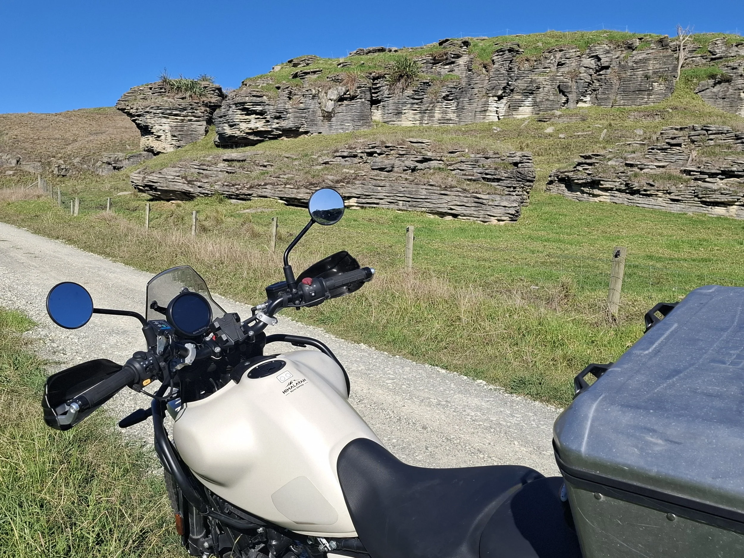 A motorcycle parked on a dirt road beside green grassy fields and rock formations under a clear blue sky.
