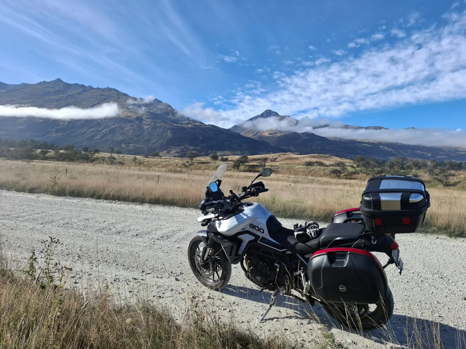 A motorcycle parked on a gravel road with mountains and a blue sky with clouds in the background.