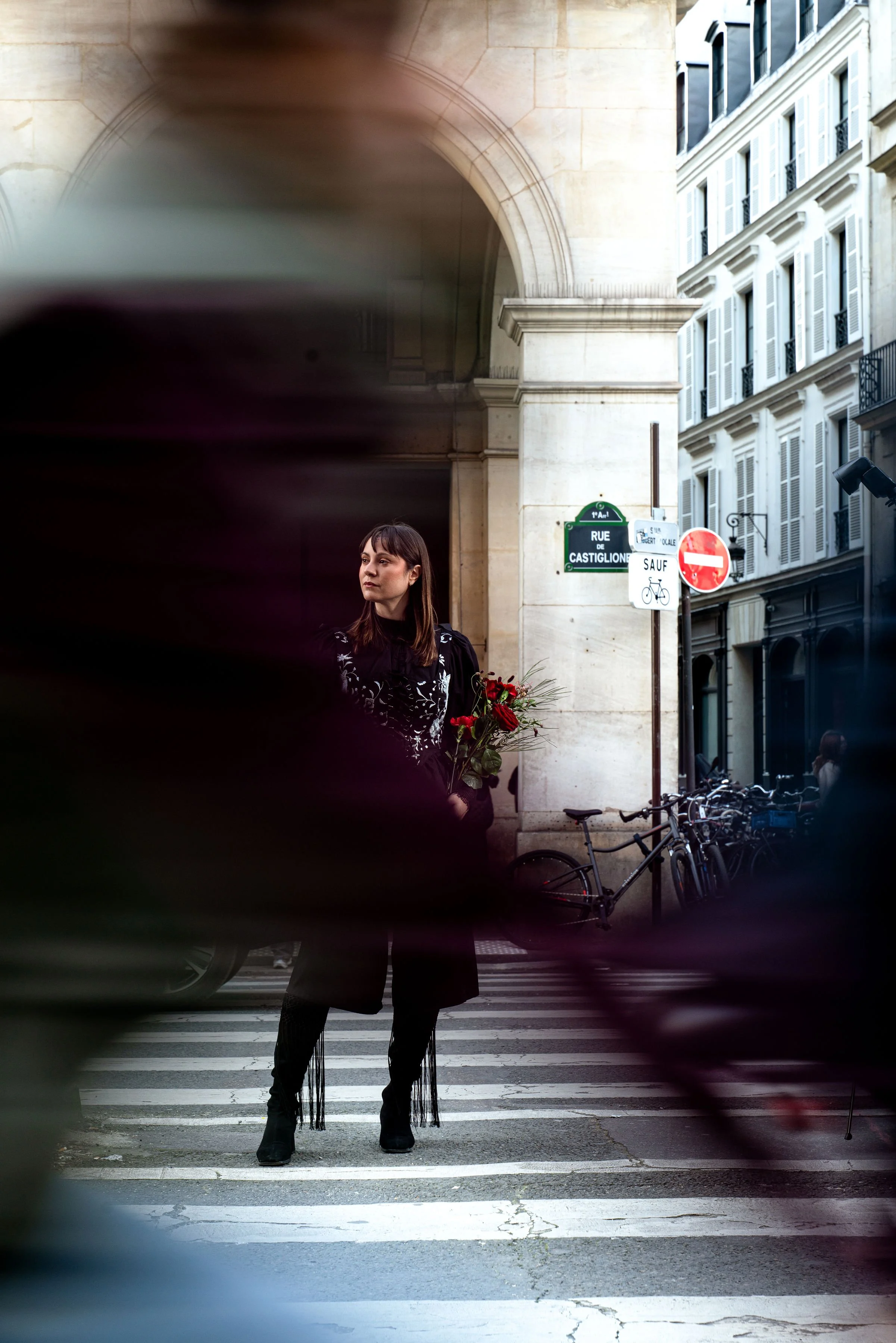 Mujer con flores en mano, vista a través de ramas de flor, en cruce peatonal en París, con edificios y señales de tráfico en el fondo.
