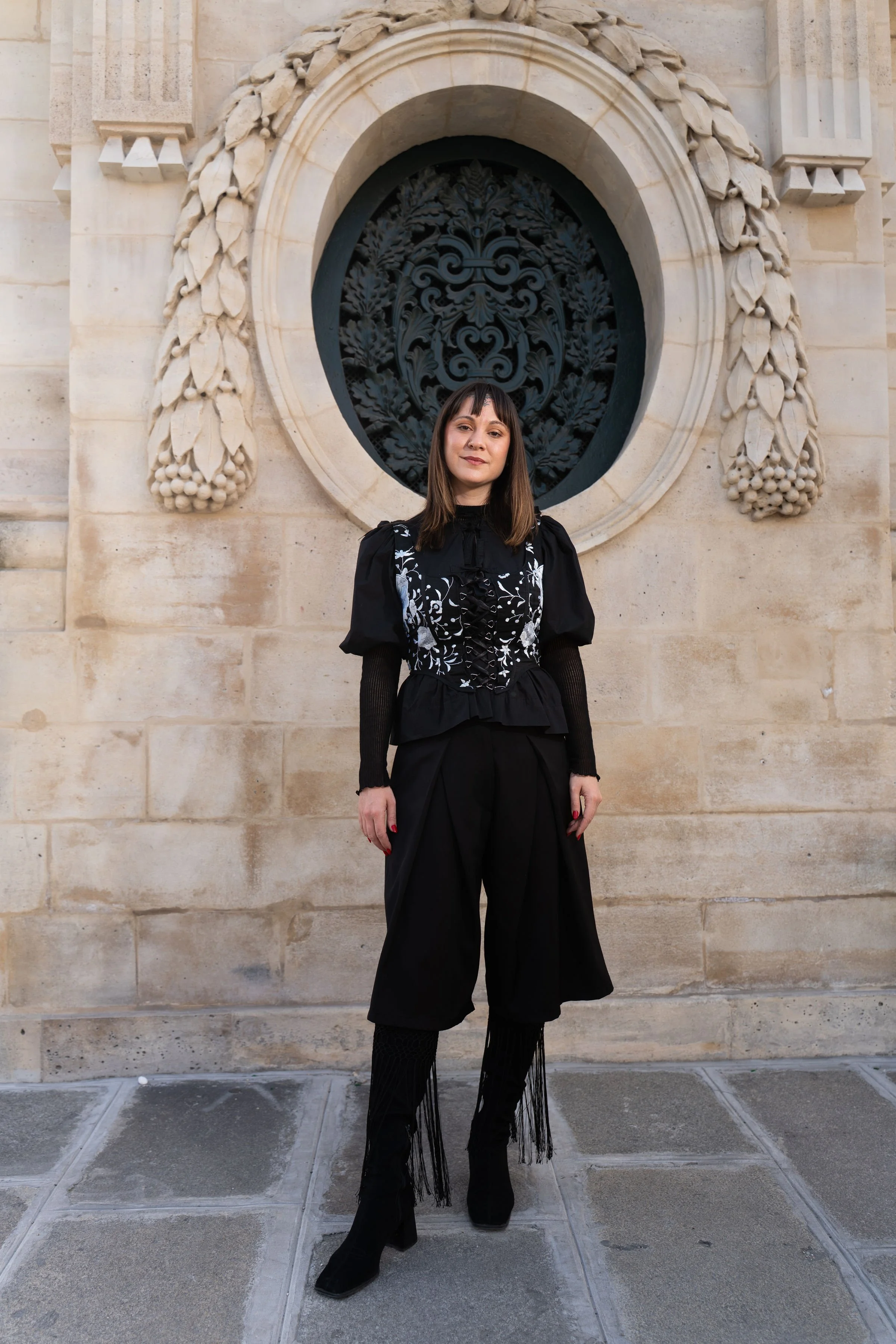 Mujer posando frente a una pared de piedra con un arco y una ventana de rejilla decorativa, vestida con ropa negra con detalles blancos.
