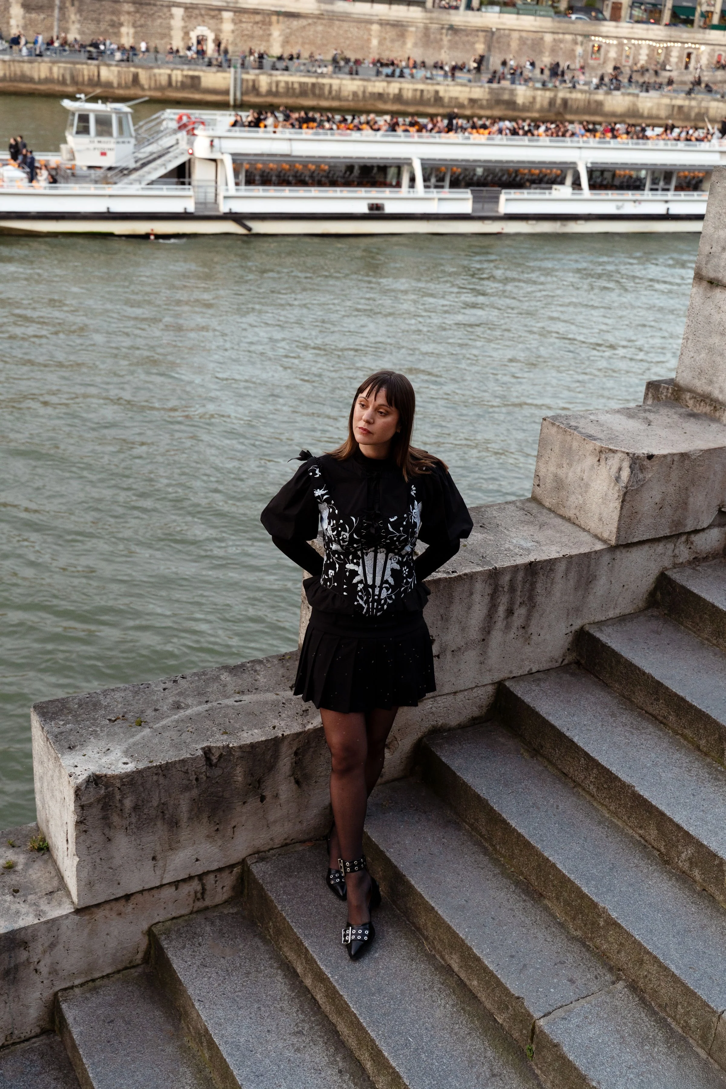 Mujer con vestido negro y tacones de charol con hebillas, posando en unas escaleras cerca del río, con un barco y un paseo lleno de gente al fondo.
