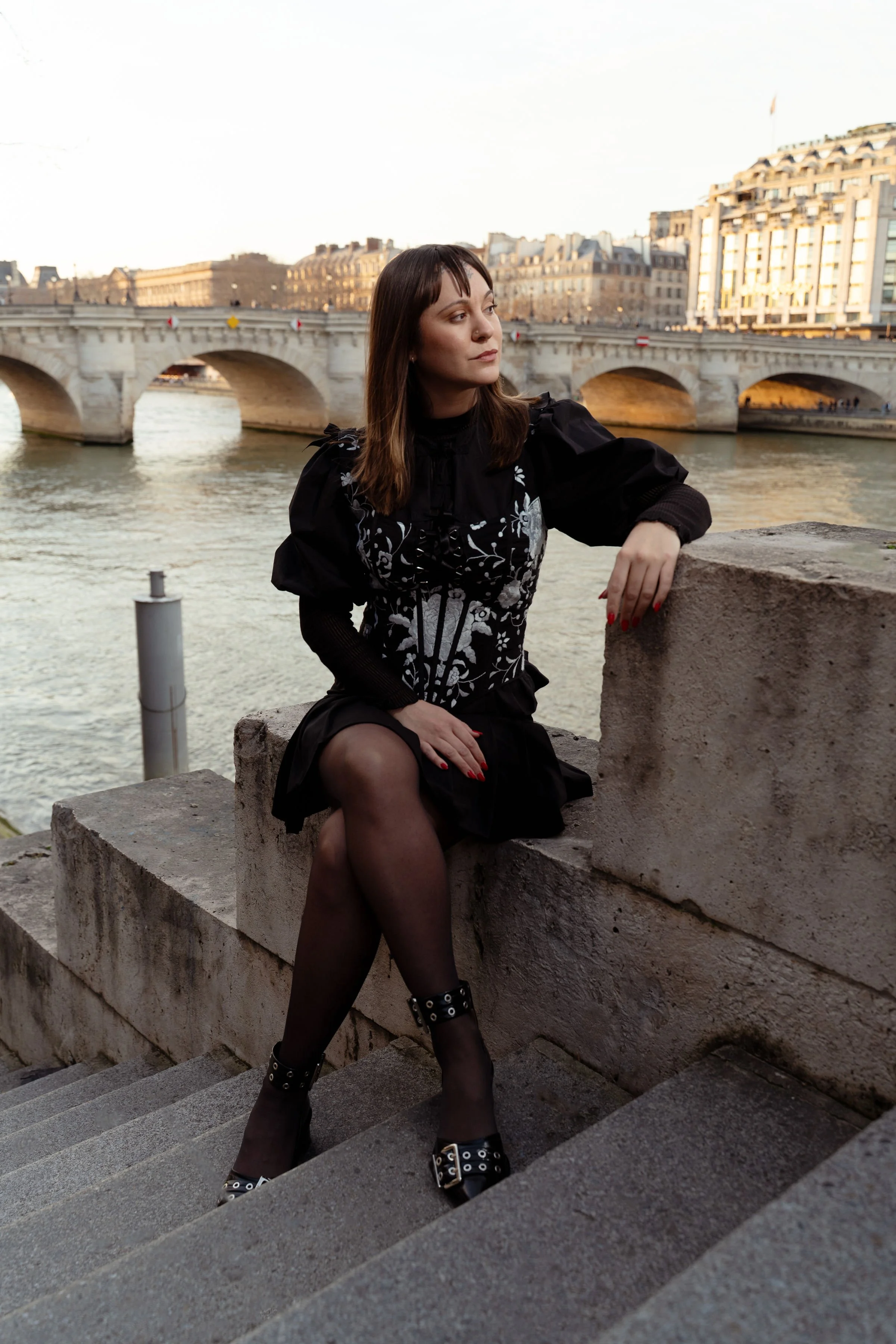 Mujer sentada en las escaleras junto al río Sena con un puente y edificios al fondo en París.