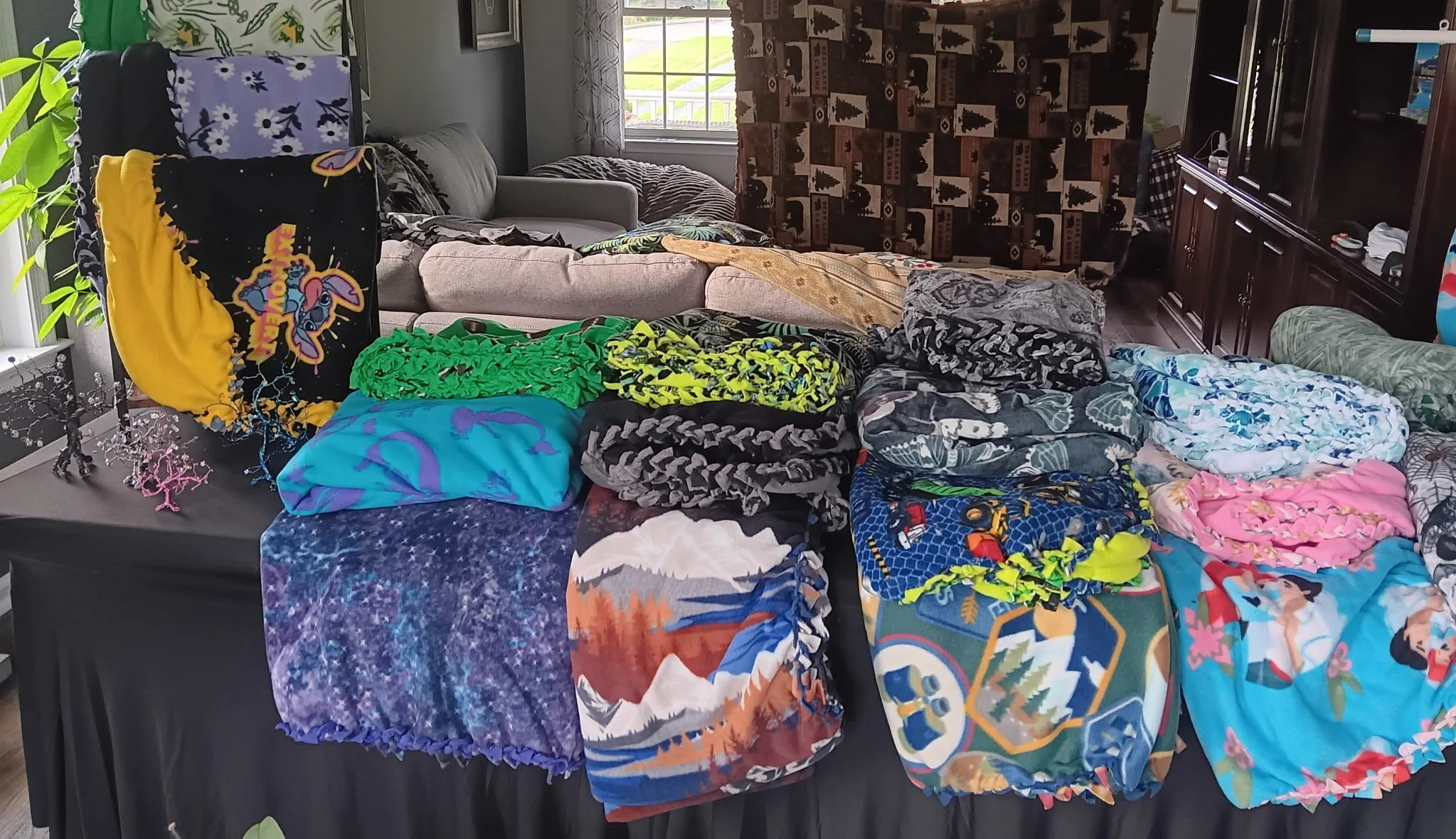 A table with various colorful blankets and scarves folded on top of it inside a living room, with couches, windows, and a wooden cabinet in the background. Fleece by Kim