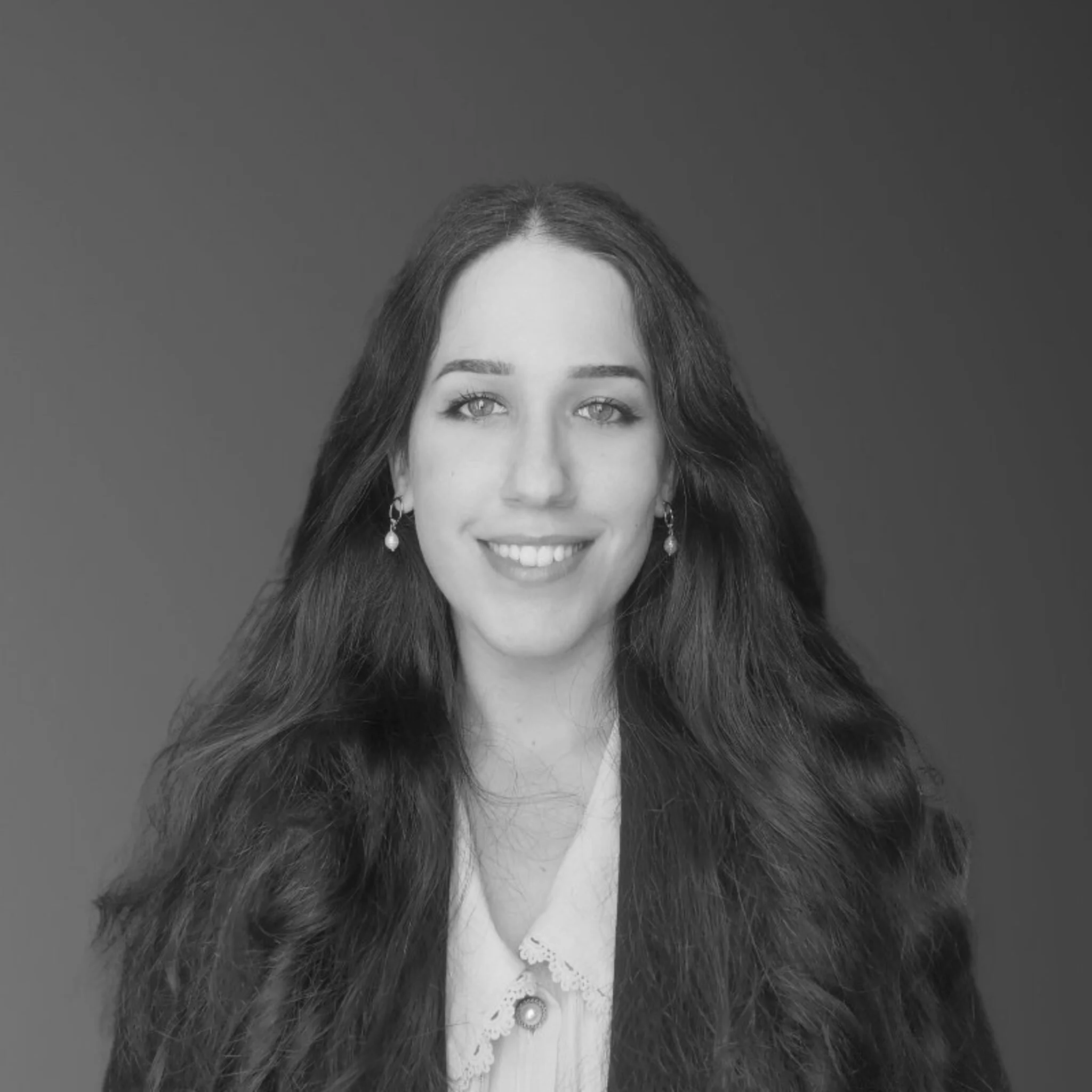Black and white portrait of a young woman with long wavy hair, earrings, and a white lace-collared shirt, smiling at the camera.