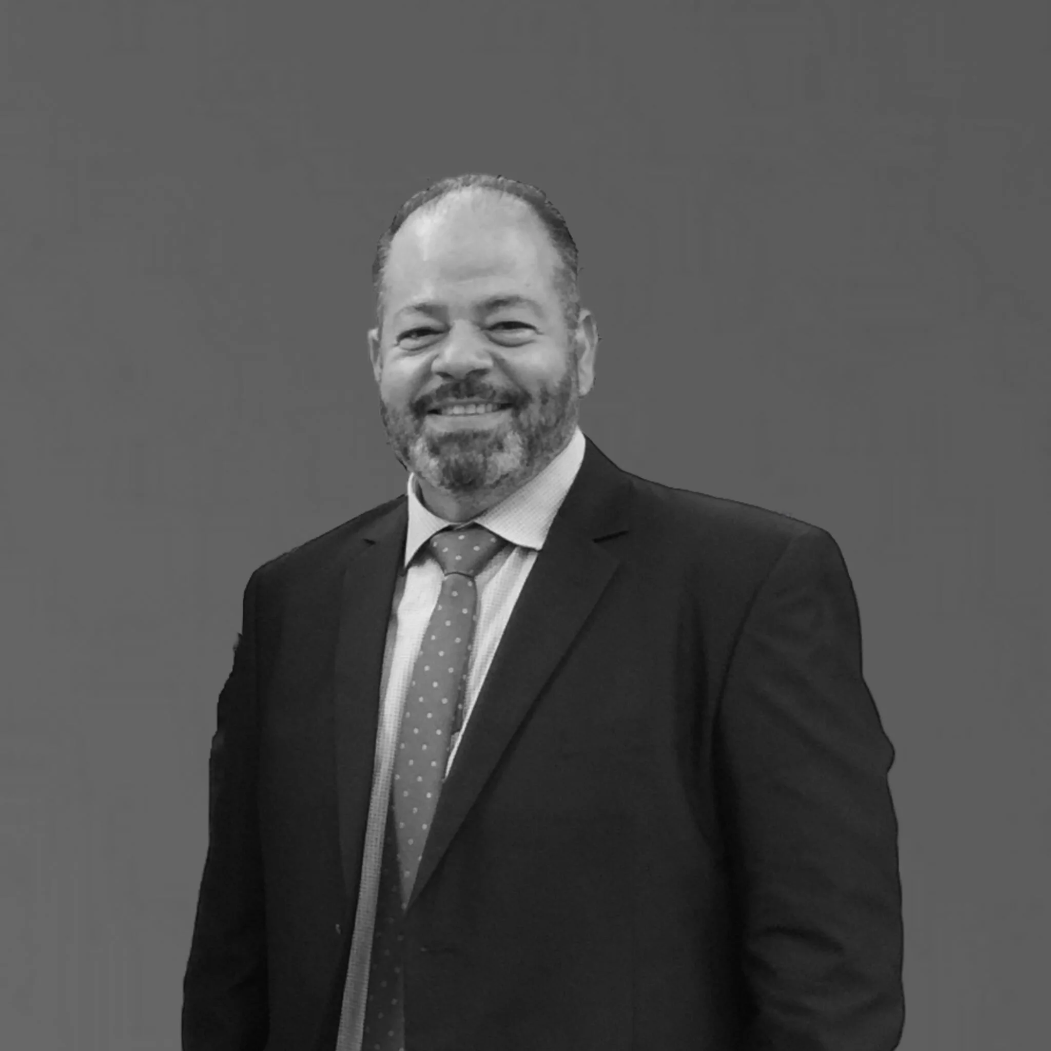 A smiling man with a beard wearing a suit, dress shirt, and tie, standing against a plain background.