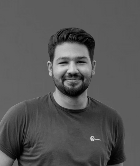 Young man with dark hair, beard, and mustache smiling, wearing a dark T-shirt with a small logo on the chest, standing against a plain gray background.