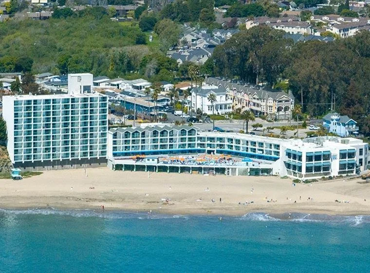 Aerial view of a beachfront hotel with a sandy beach in front, next to the ocean. The hotel has multiple stories with balconies, and there are palm trees and other buildings in the background.