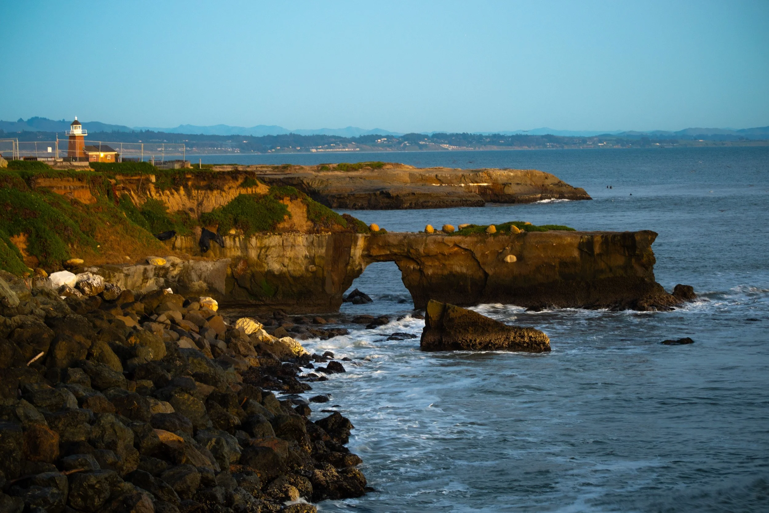 Sunset view of a rocky coastline with a natural arch, green vegetation, and a lighthouse in the distance.