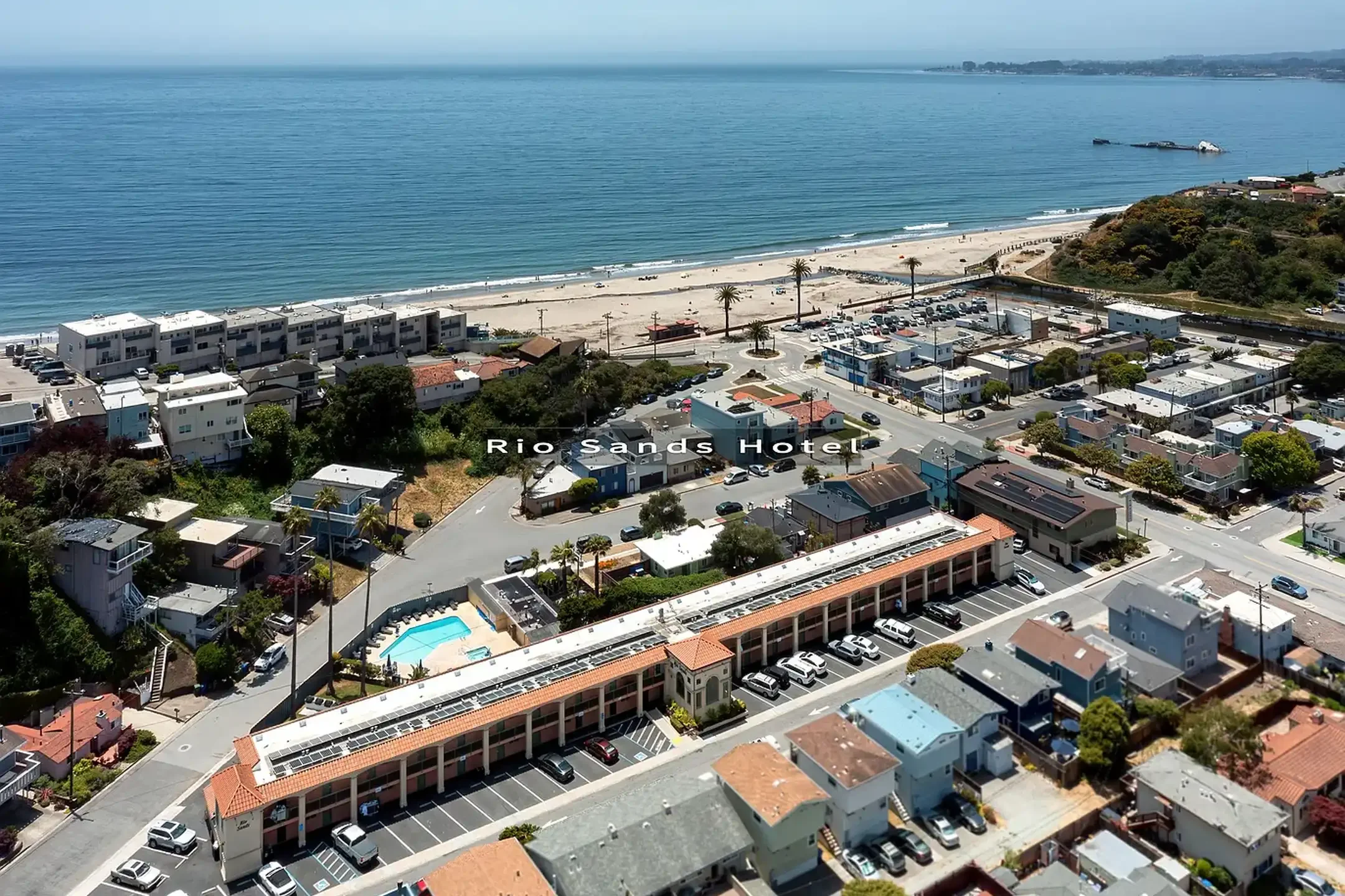 Aerial view of Rio Sands Hotel, a beachfront property with a parking lot, residential houses, and a sandy beach with palm trees along the coast.