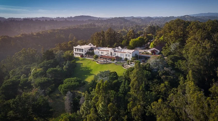 A large, luxurious house surrounded by lush green trees on a hillside, with a spacious lawn and outdoor patio area, under a clear blue sky.