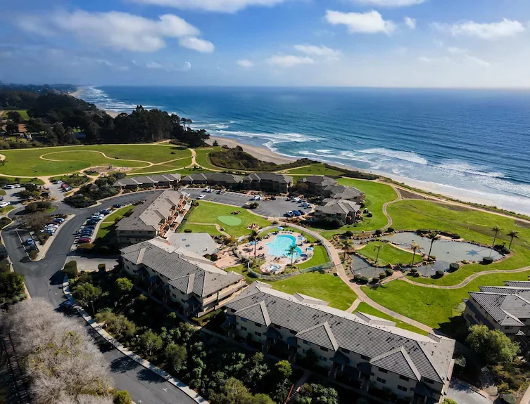 Aerial view of a beachfront resort with swimming pools, green lawns, walking paths, nearby parking lot, and the ocean in the background.