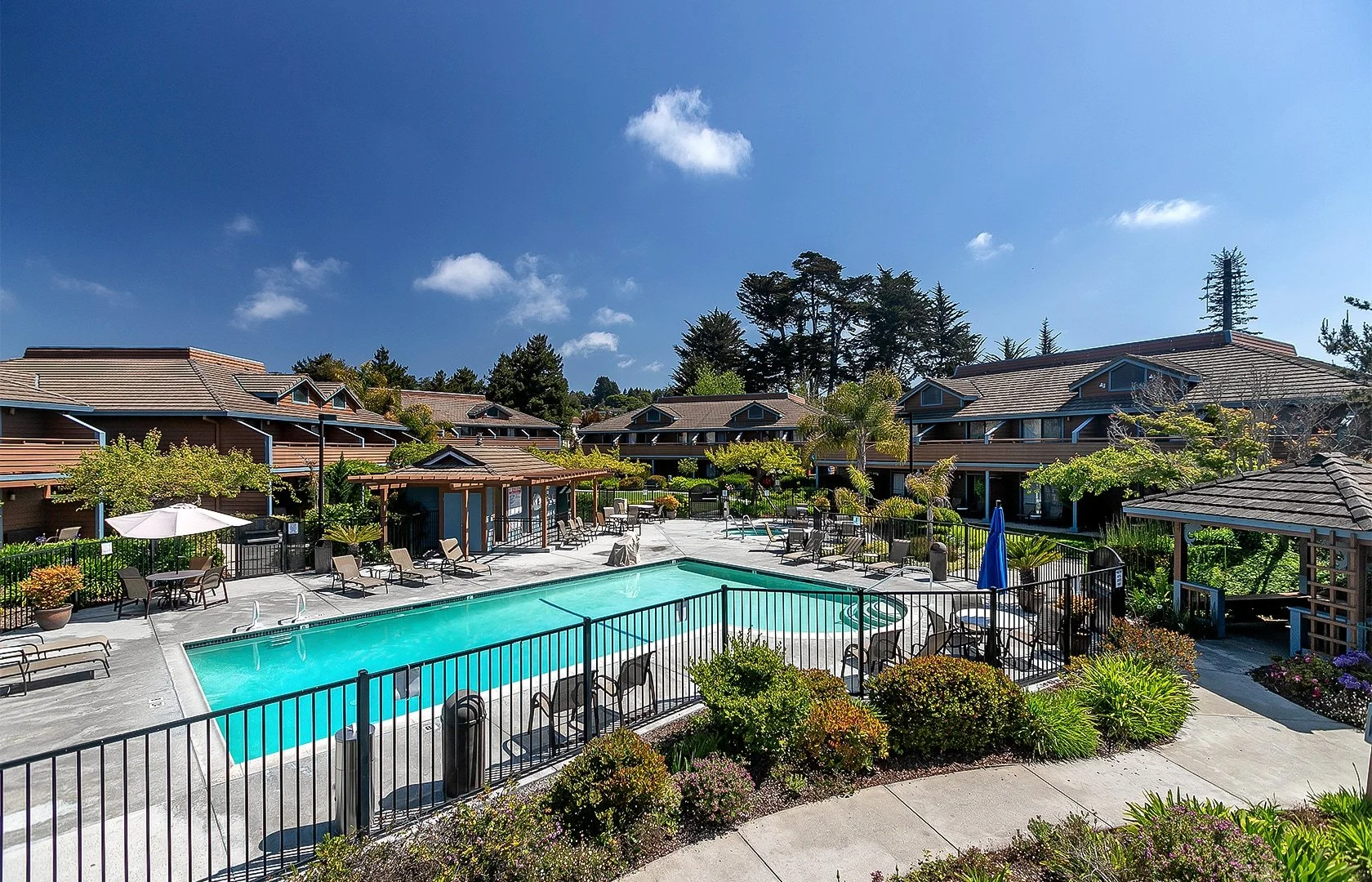 Outdoor swimming pool area in a residential complex with lounge chairs, umbrellas, trees, and multi-story buildings in the background under a sunny blue sky.