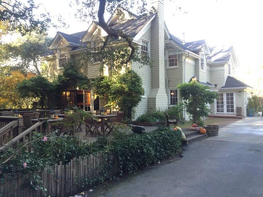 A large residential house with beige siding and multiple gabled roofs. It has several windows and a patio area with wooden outdoor furniture, pumpkins, and decorative plants. There are trees and shrubs surrounding the house, creating a cozy and inviting atmosphere.