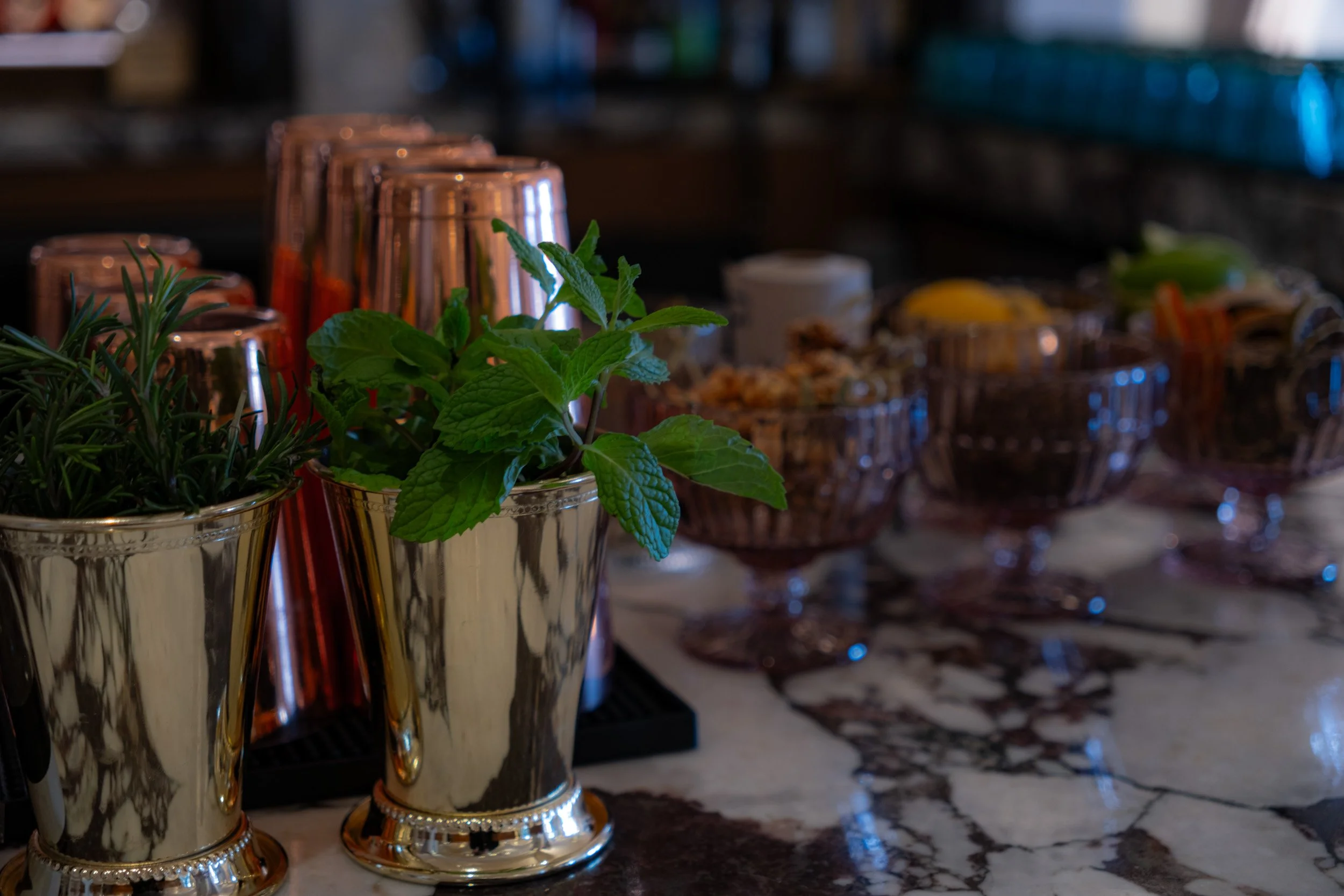 Mint and rosemary herbs in silver cups, with pink cocktail glasses and bowls of snacks in the background, on a marble countertop.