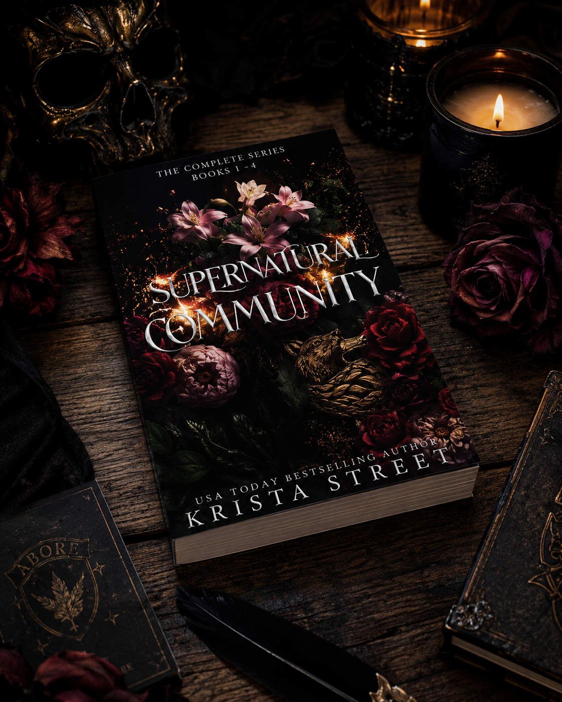 A book titled 'Supernatural Community' by Krista Street is on a dark wooden table surrounded by candles, flowers, a skull decoration, and gothic accessories in a dimly lit setting.