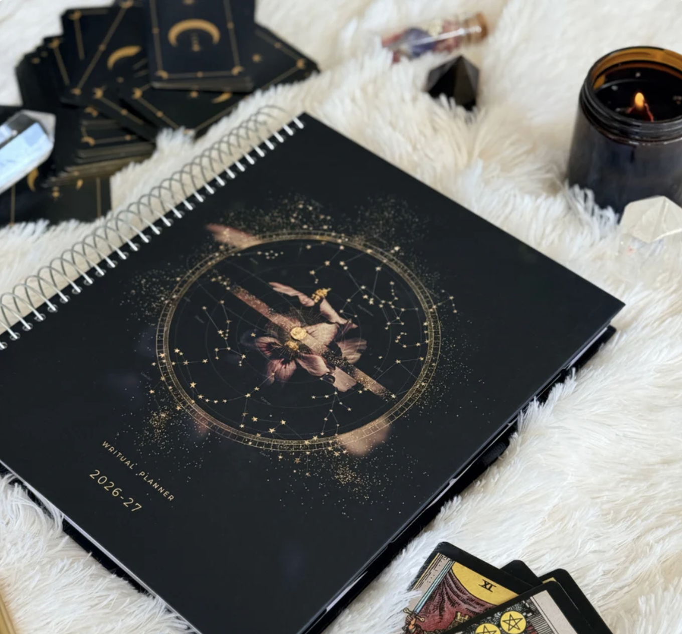 A black spiral-bound notebook with gold and pink celestial and floral design on the cover, labeled as a spiritual or mystical planner for 2026-27, resting on a white furry surface. Surrounding items include tarot cards, a lit candle in a dark jar, and crystals.