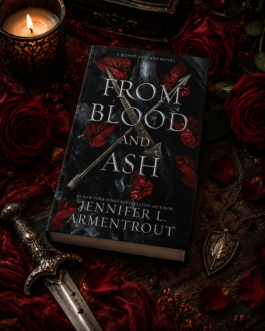 Close-up of a book titled "From Blood and Ash" by Jennifer L. Armentrout, surrounded by red roses, a lit candle, a sword with a detailed hilt, and a necklace with a leaf pendant on a dark wooden surface with rose petals.