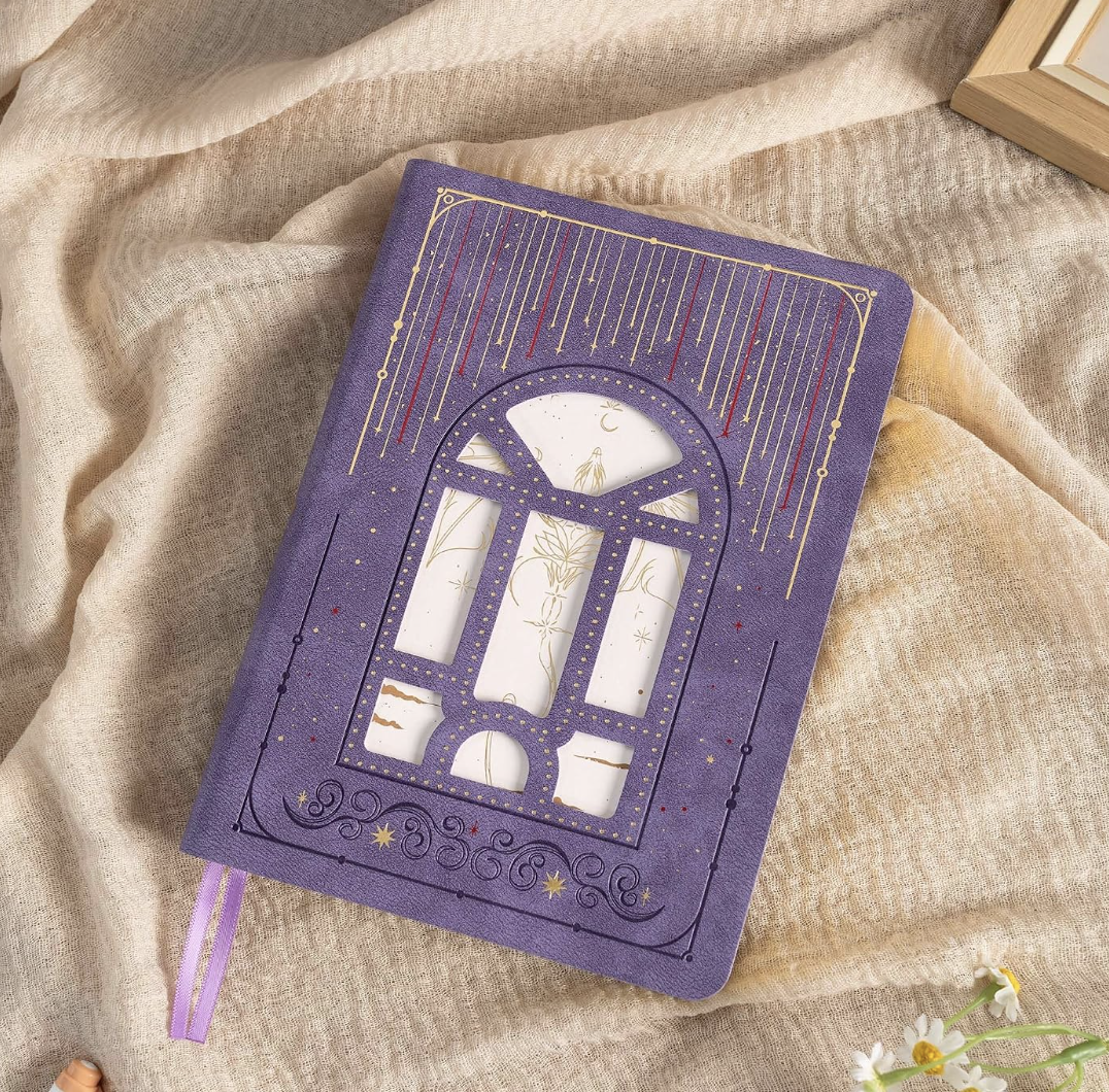 A purple hardcover notebook with star, moon, and constellation designs, featuring a window-like pattern on the cover, placed on beige fabric next to a wooden frame and small flowers.