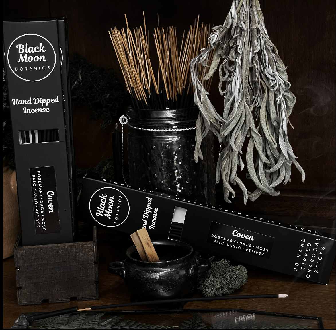 A collection of Black Moon Botanics incense products, including hand-dipped incense boxes labeled Covan and a jar of incense sticks, arranged with dried lavender, sage, and moss on a wooden surface.