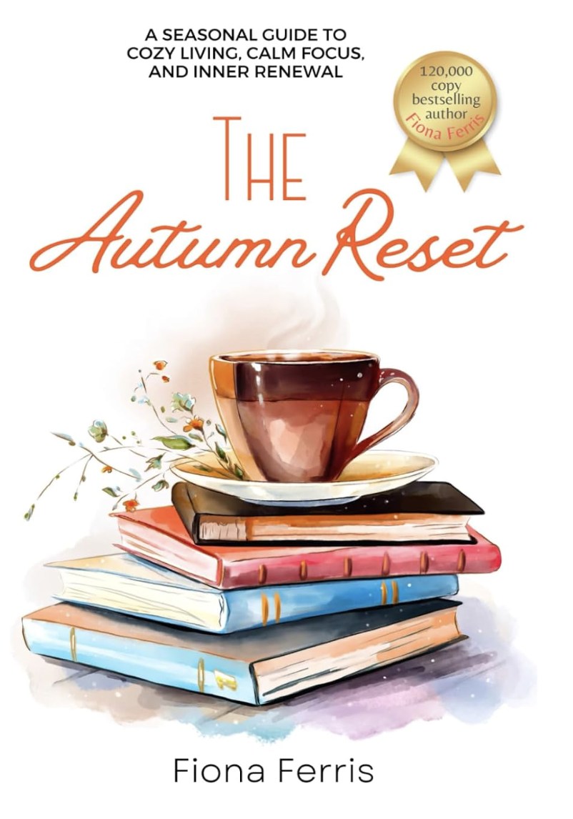 Book cover titled "The Autumn Reset" with the subtitle "A seasonal guide to cozy living, calm focus, and inner renewal" by Fiona Ferris. The cover features a steaming cup of tea on top of a stack of five colorful books with a small sprig of flowers beside it.