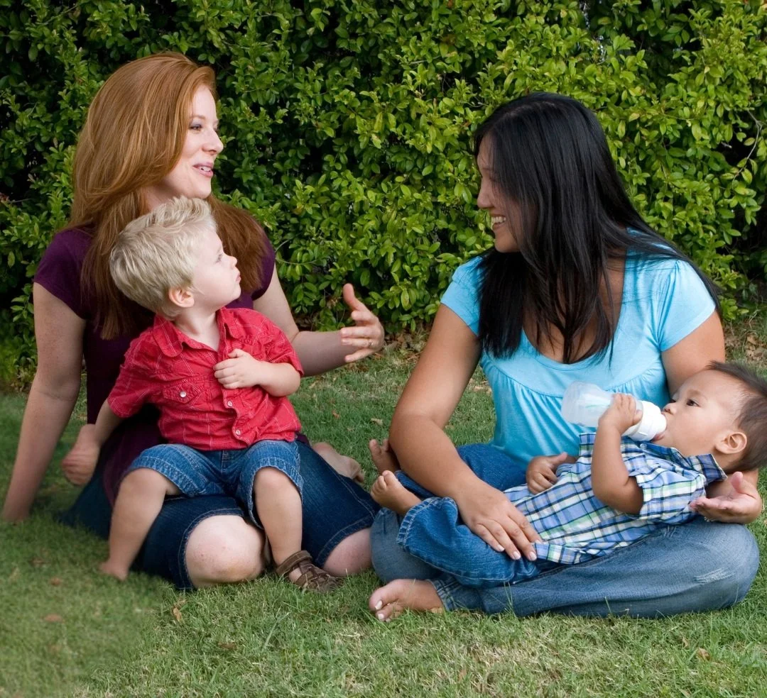 Two women sitting outdoors on grass, each with a young child, smiling and engaged in conversation, with green bushes in the background.
