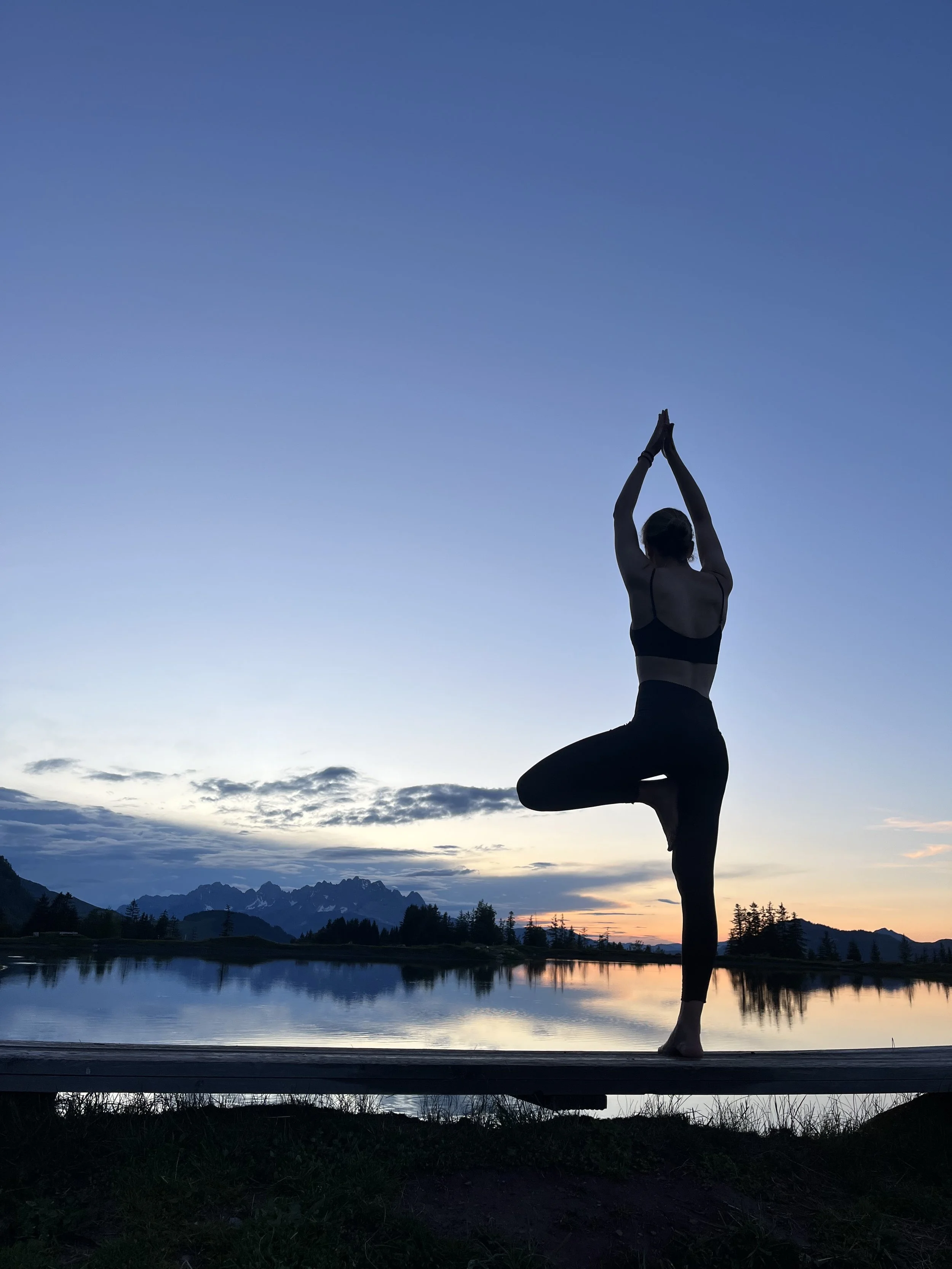 Eine silhouette einer Person, die Yoga im Lotussitz bei Sonnenuntergang am See macht, mit Bergen im Hintergrund.