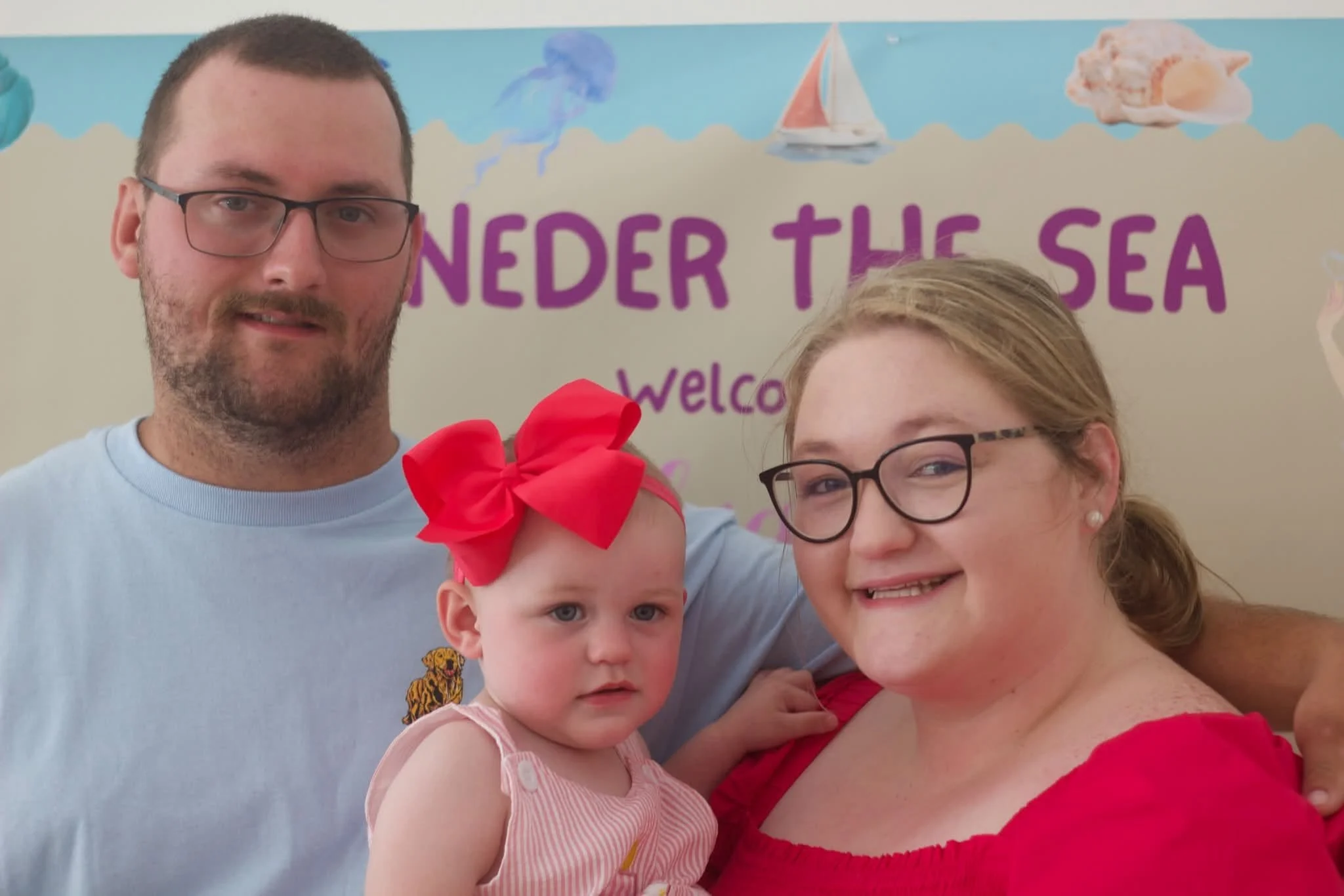 A family of four taking a selfie in front of a sea-themed background that reads 'Under the Sea'. The man has glasses and a beard, the woman has glasses and blonde hair, the older girl has light brown hair and glasses, and the younger girl has a large pink bow in her hair and is wearing a pink and white sleeveless dress.