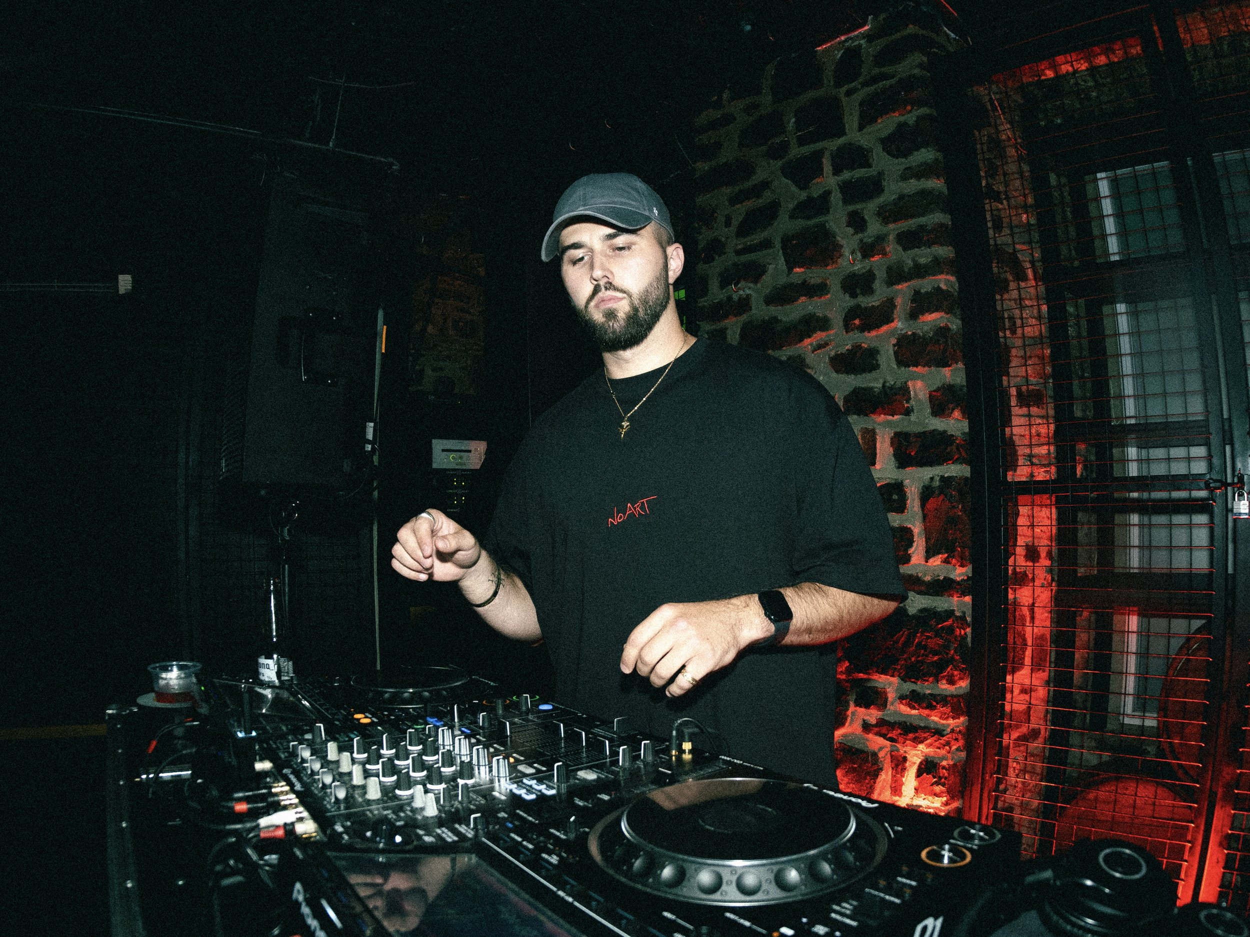 A DJ with a beard, wearing a black t-shirt, gray cap, and smartwatch, performs in a dark, industrial-style club with a brick wall and metal bars behind him, illuminated by red lighting.
