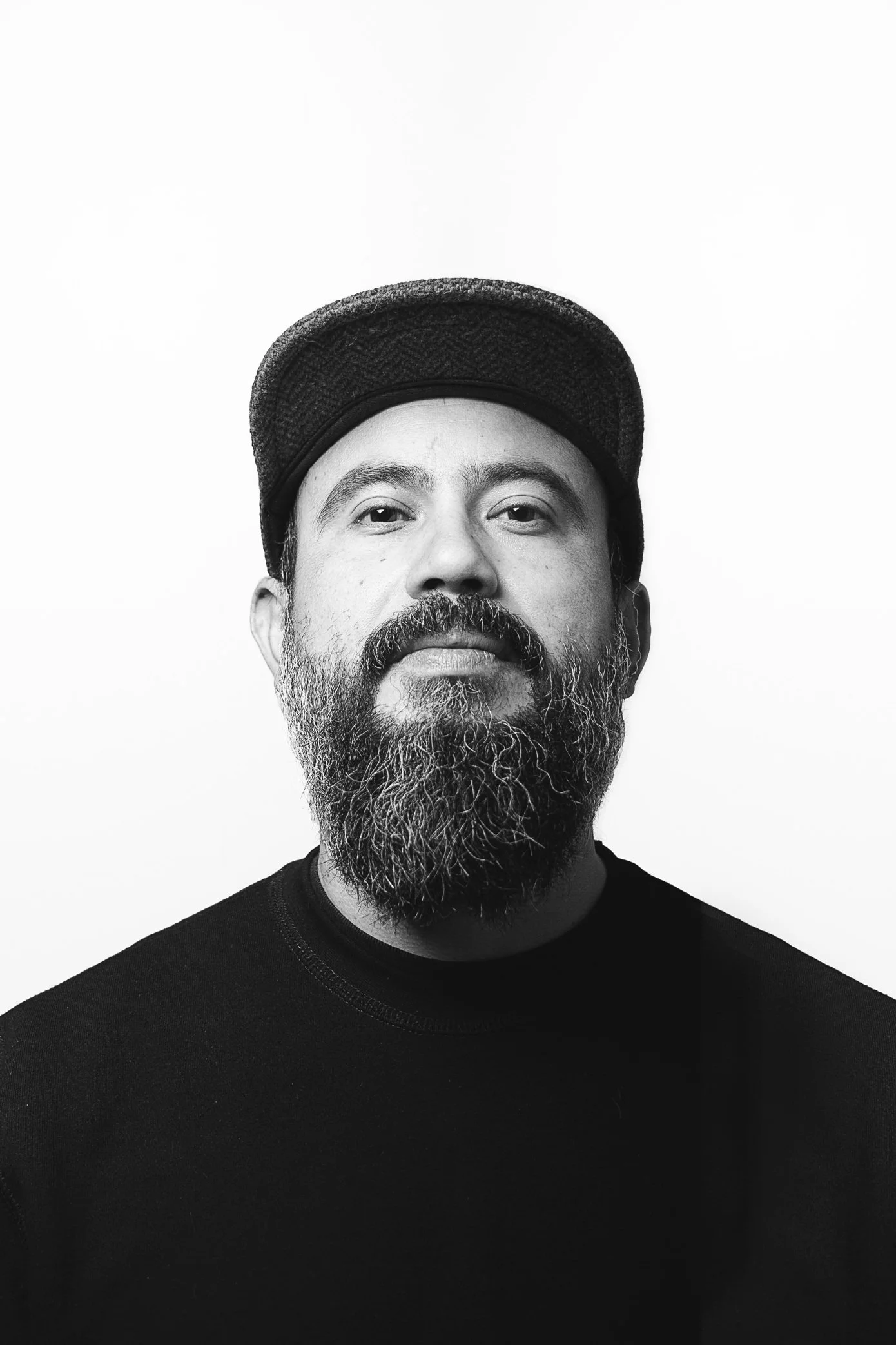 Black and white portrait of a man with a beard, wearing a cap and a dark shirt, against a plain white background.
