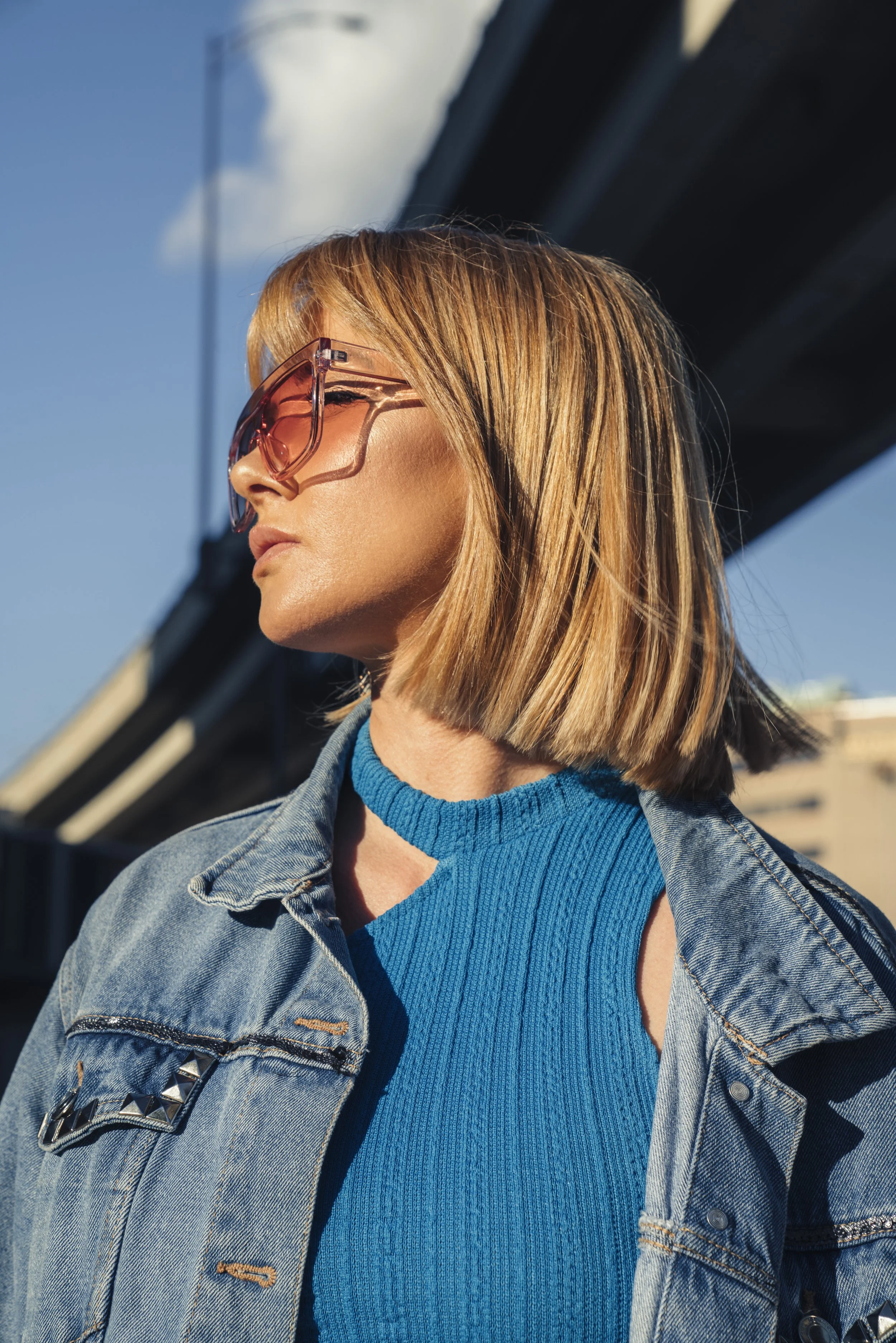 A woman with shoulder-length blonde hair wearing large pink sunglasses, a bright blue sleeveless knit top, and a denim jacket with metal stud details, standing outdoors under a bridge with a blue sky and some clouds.