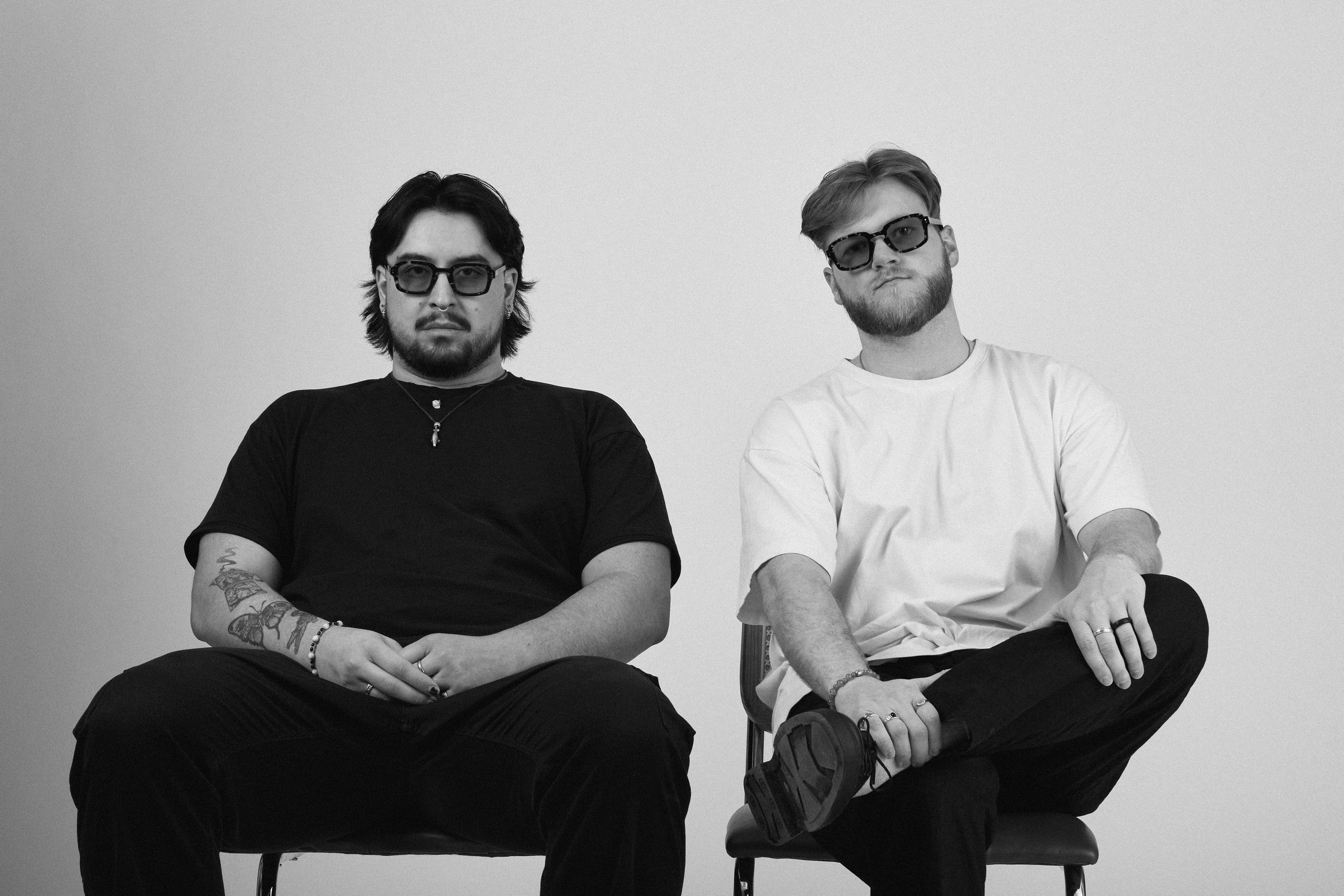 Two young men with facial hair and sunglasses sitting on chairs against a plain white wall, one with long dark hair, tattoos and a black shirt, the other with short blond hair, a white shirt, and crossed legs.