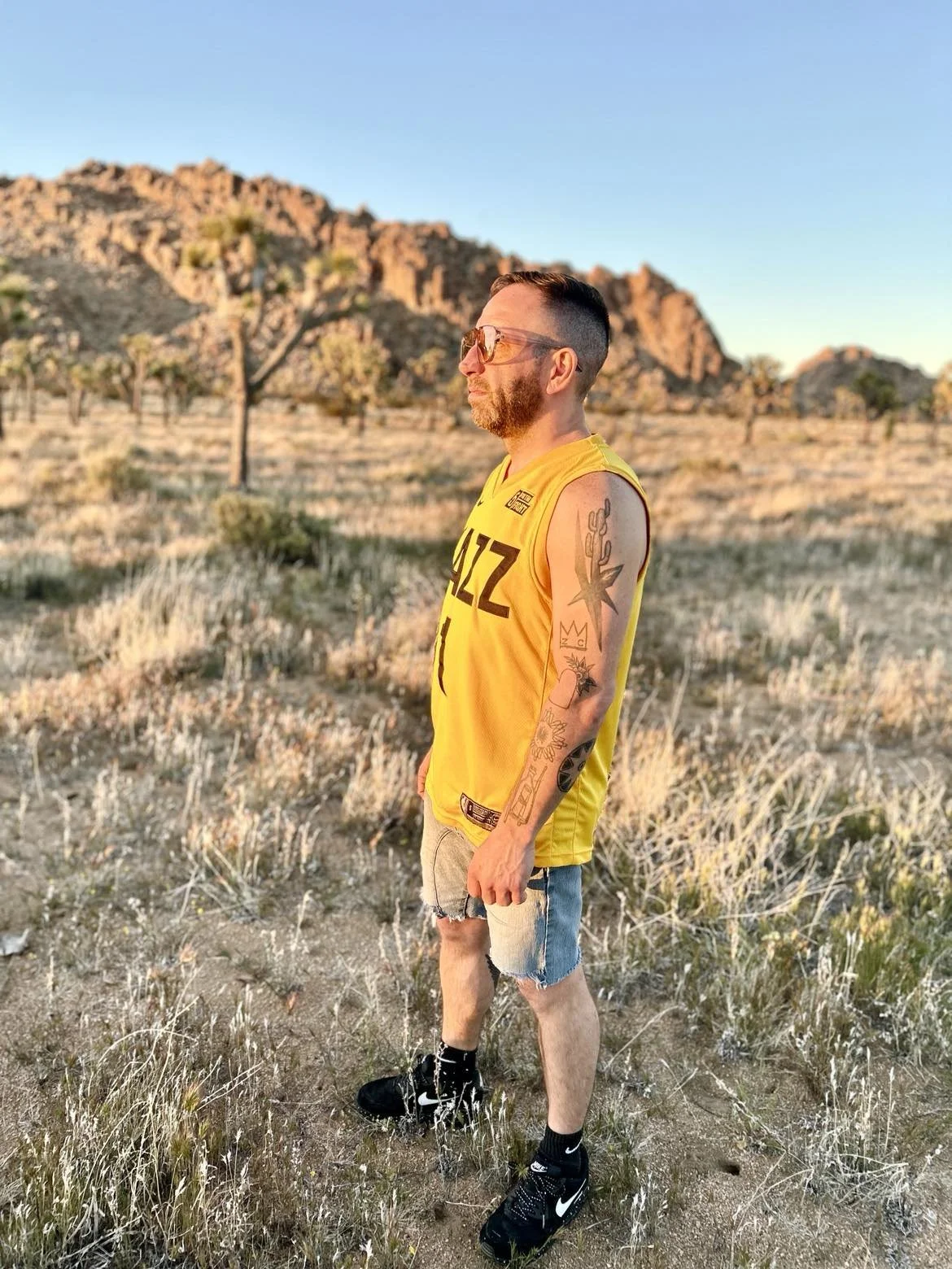 A man with tattoos, wearing a yellow sports jersey, khaki shorts, and black sneakers, standing in a desert landscape with rocky hills and sparse trees during sunset.