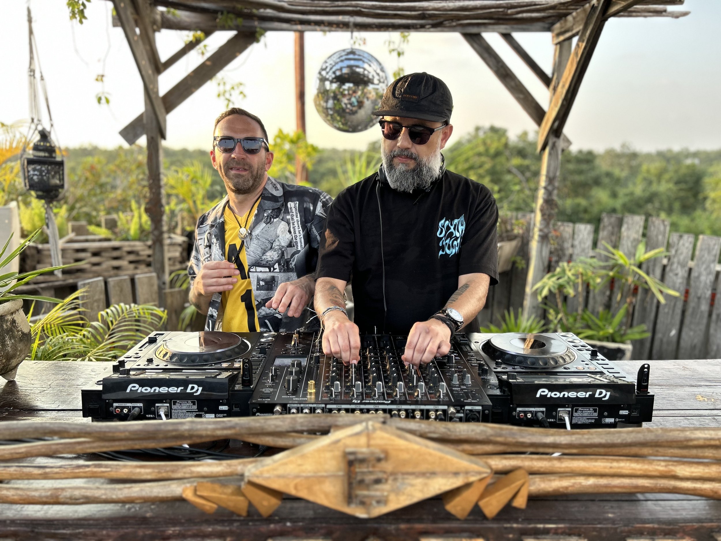 Two men with DJ equipment outdoors; one wearing sunglasses and a patterned shirt, the other with a beard, sunglasses, and headphones, operating a DJ mixer in a rustic setting.