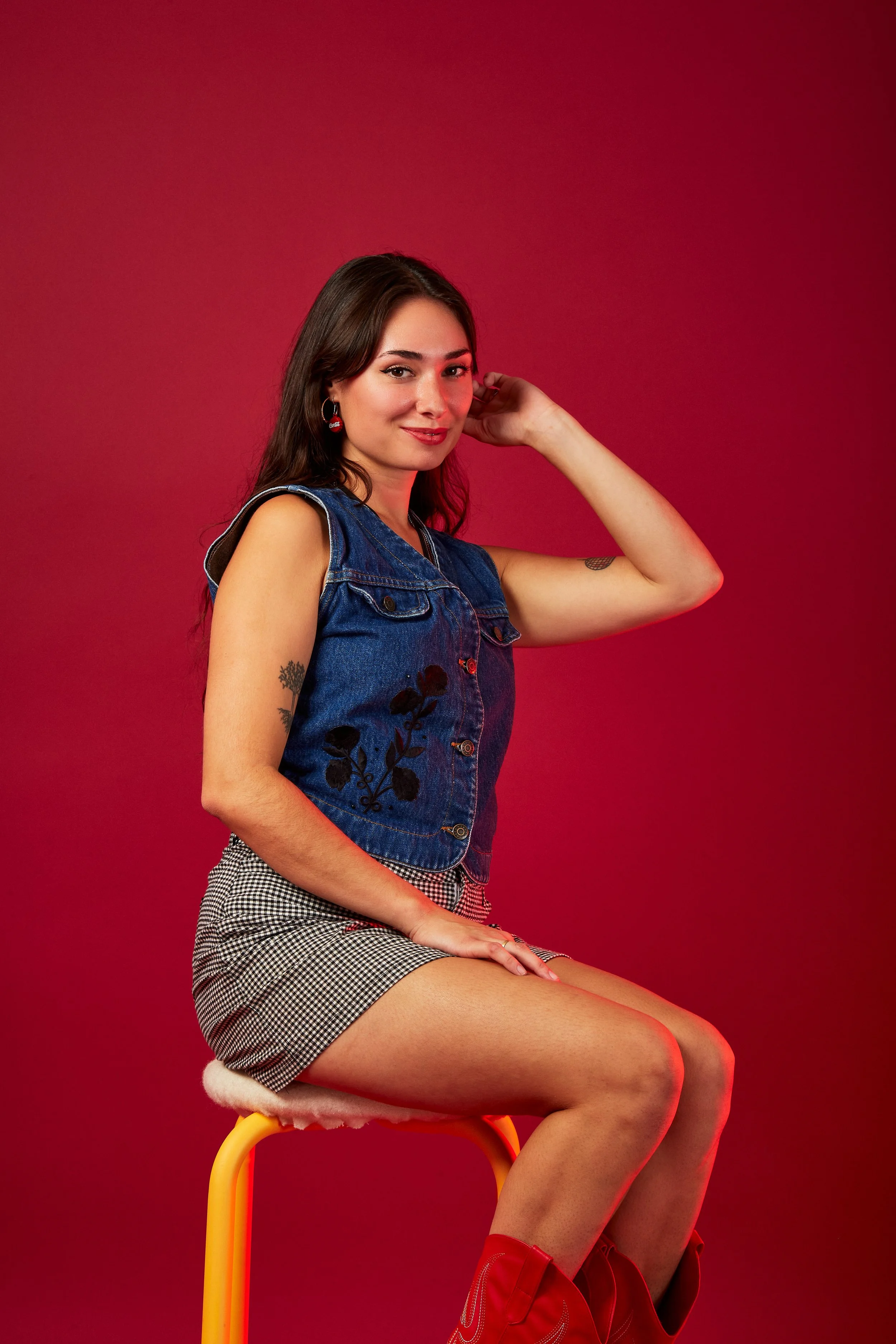 A young woman with dark brown hair sitting on a yellow stool against a red background, wearing a sleeveless denim vest, black and white checkered shorts, and red boots, with tattoos on her arms and red earrings.