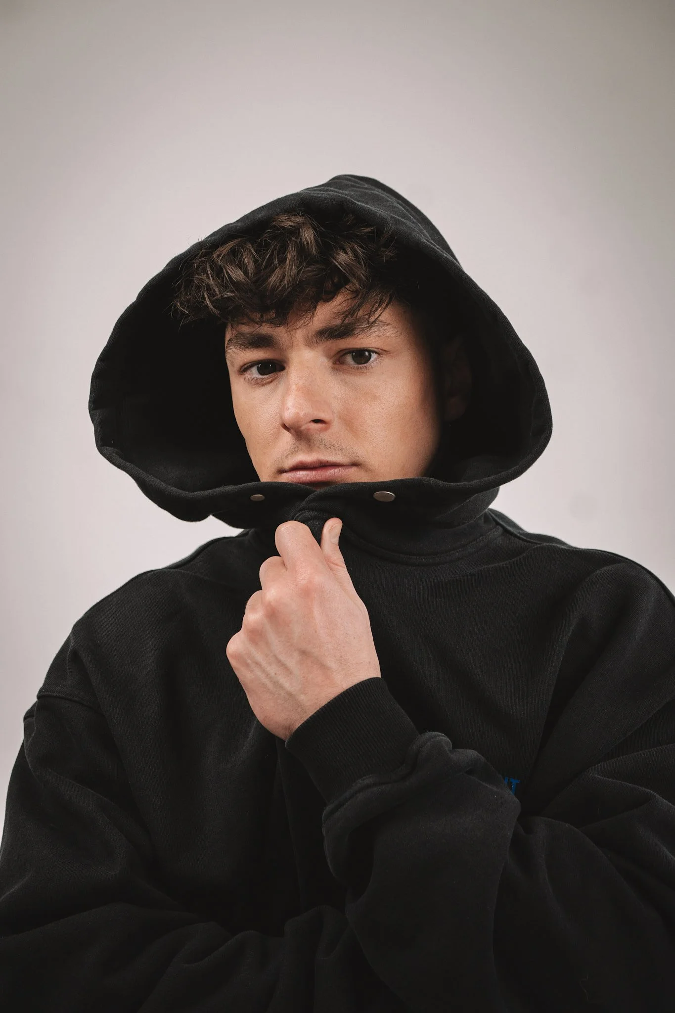 Young man wearing a black hoodie, pulling the hood up with one hand, looking directly at the camera with a neutral expression.