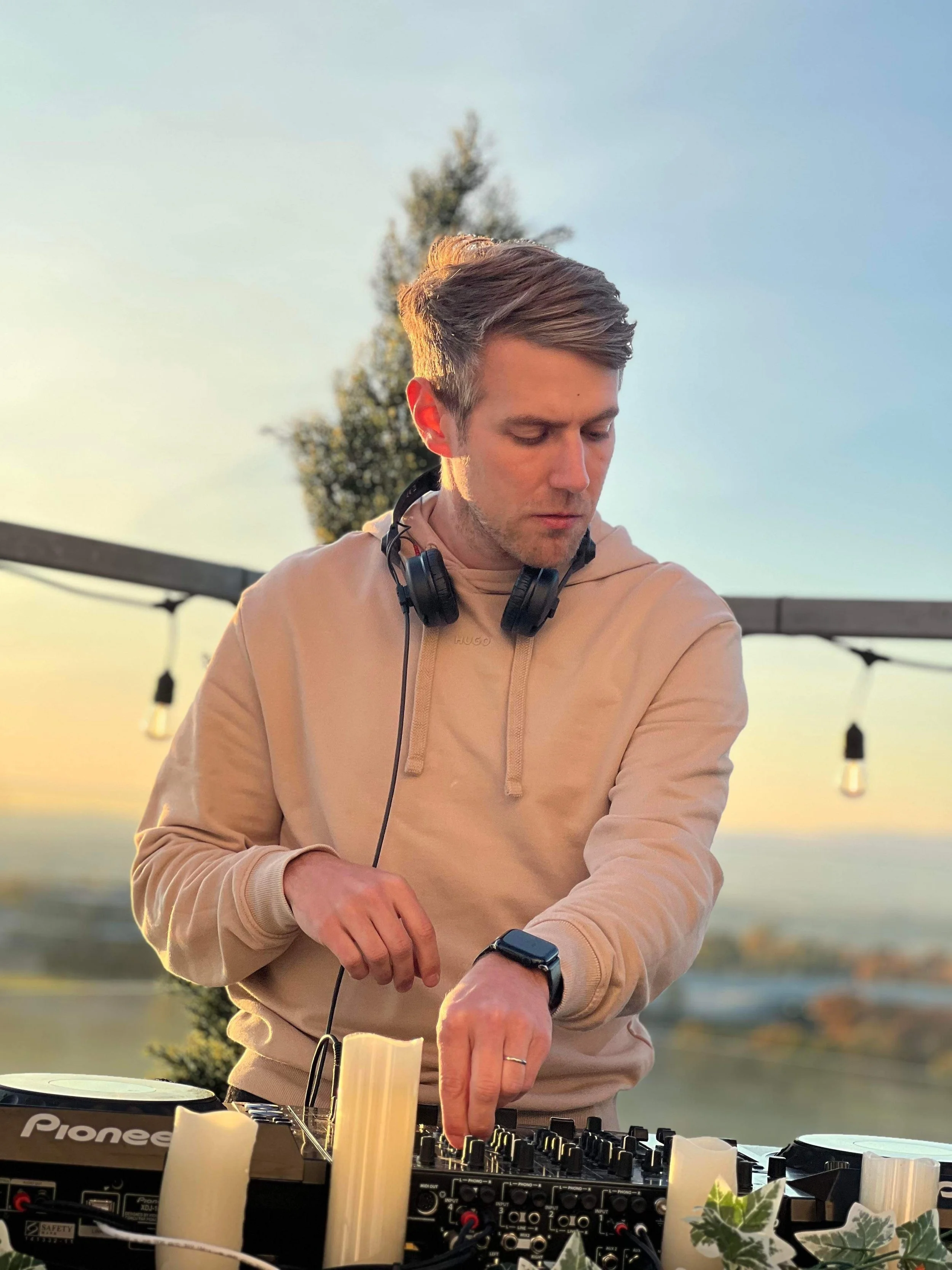 A male DJ with headphones around his neck, wearing a beige hoodie, is adjusting equipment at an outdoor event during sunset. There are decorative candles and plants on the table, with string lights hanging in the background.